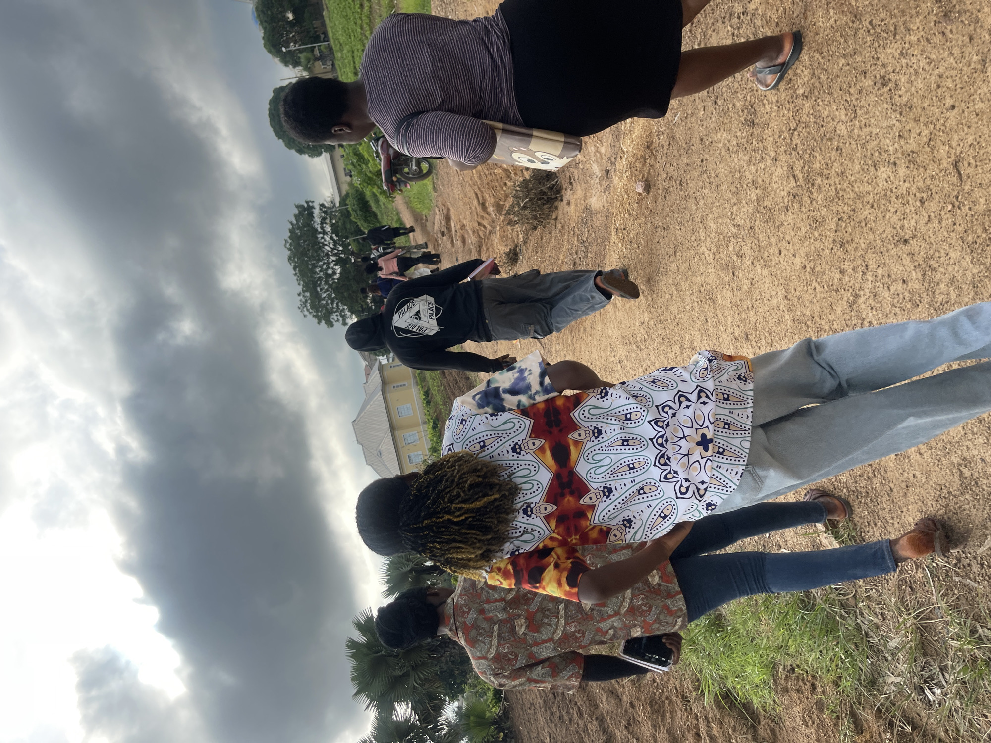 The image depicts a group of people walking on a dirt path in a rural or semi-rural area. The sky is overcast with dramatic clouds, suggesting a potential for rain. In the background, there are buildings, including a light-colored house with a yellow roof, and vegetation, including trees and what appears to be a cultivated field on the right. A motorcycle is parked on the right side of the path. The people are walking away from the camera. The person closest to the camera on the right is wearing a black skirt and a striped shirt, carrying a patterned tote bag. To their left, a person in the foreground is wearing a brightly patterned shirt and light-colored pants. Further down the path, other individuals are visible, including one in a black hoodie and cargo pants. The ground is a mix of dirt and sparse vegetation. The overall impression is of a casual outdoor gathering or movement within a community. The location is identified as Rafin Par, Nigeria.