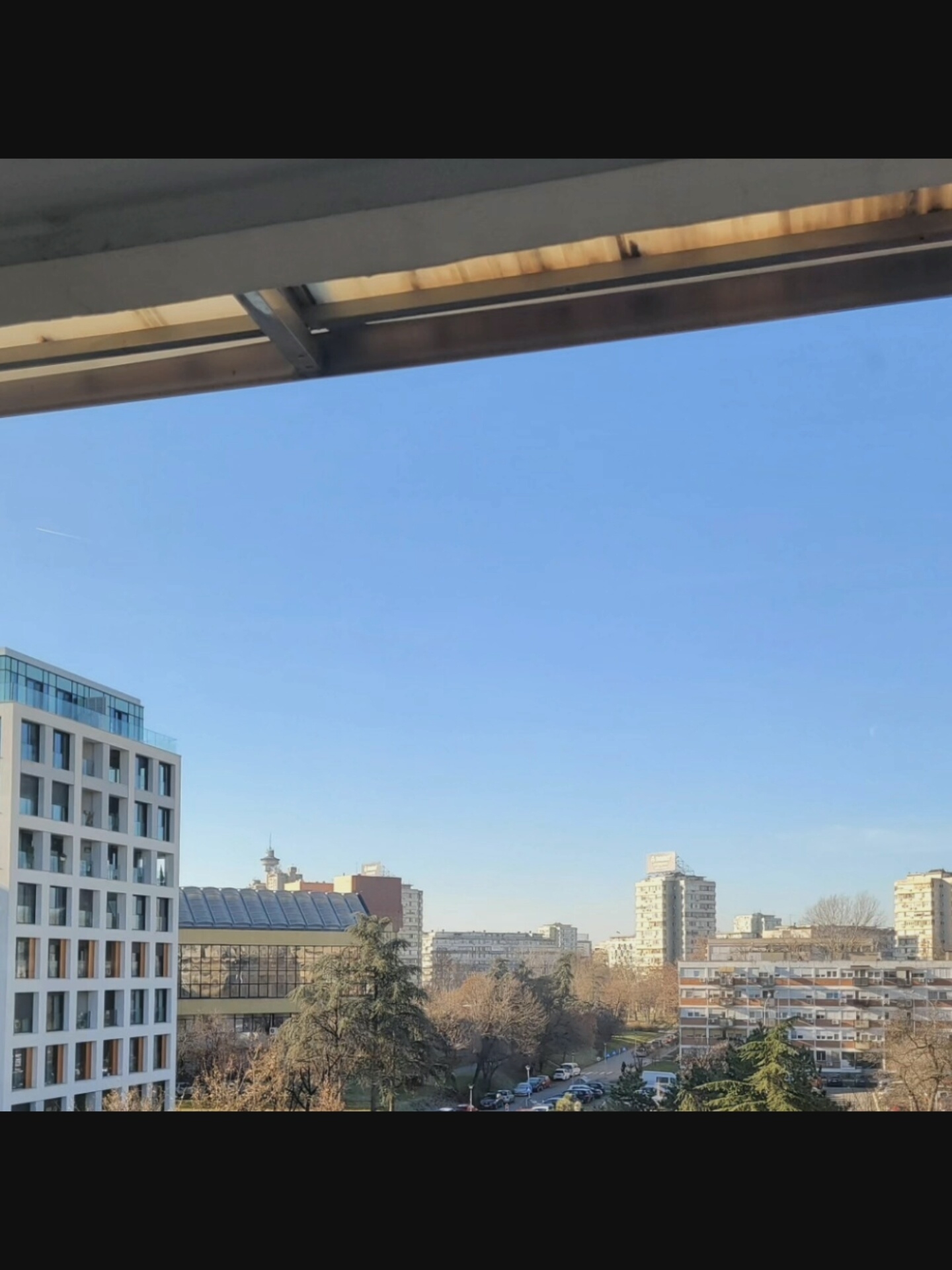 The image is an outdoor, daytime shot taken from an elevated position, likely a balcony or window, looking out over a cityscape. The foreground features the underside of a structure, possibly an awning or roof overhang, with visible wooden slats and metal supports.
The main scene depicts a clear, bright blue sky with a faint contrail from an aircraft. Below the sky, a modern white building with numerous glass-paneled windows and balconies dominates the left side of the frame. To the right of this building, a longer structure with a reflective, glass-covered facade and a sloped roof is visible. Further into the background, trees with bare branches are interspersed with apartment buildings of varying architectural styles and heights. Some buildings are tall and slender, while others are wider and appear to be older blocks of flats with numerous small windows. A street with parked cars is visible between the trees and buildings. The overall impression is an urban environment during the day, likely in a temperate climate during the late autumn or winter due to the bare trees.