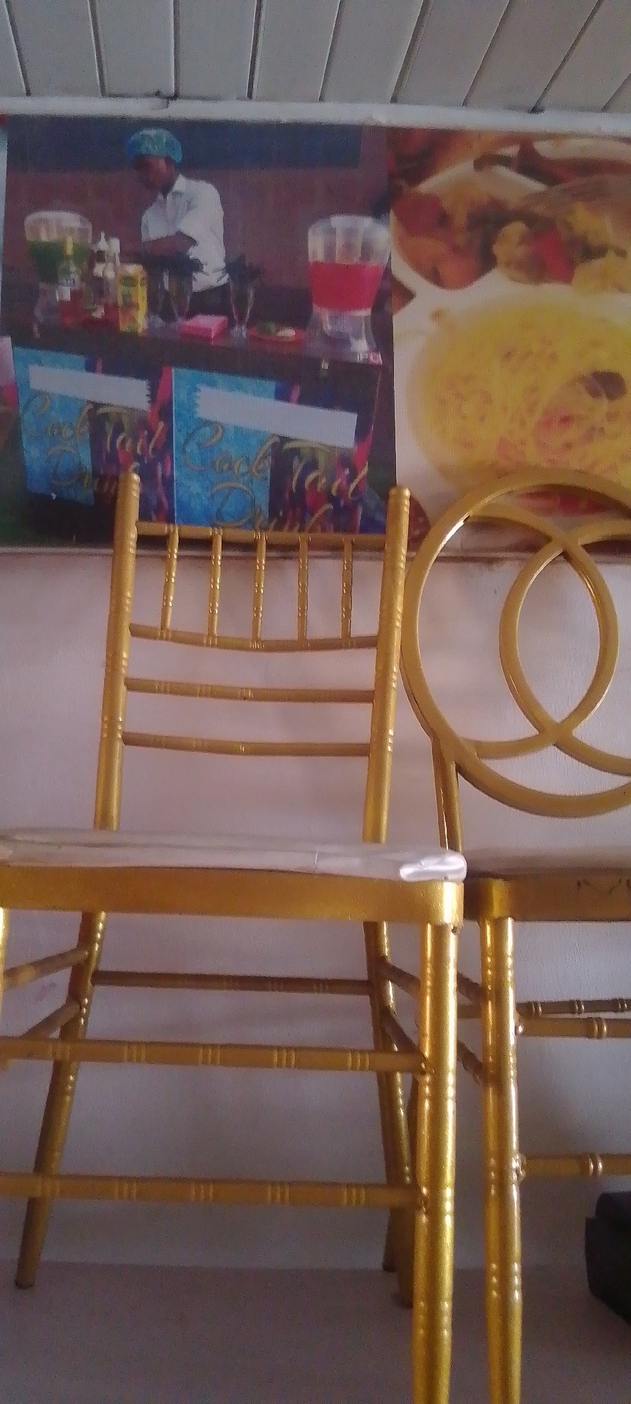 The image depicts two gold-colored chairs positioned in front of a wall. A photograph is mounted on the wall behind the chairs, showing a person wearing a blue hairnet and a white shirt preparing drinks and a close-up of food. The setting appears to be indoors, possibly in the city of Unknown, Unknown, but lacks specific details for precise location identification.