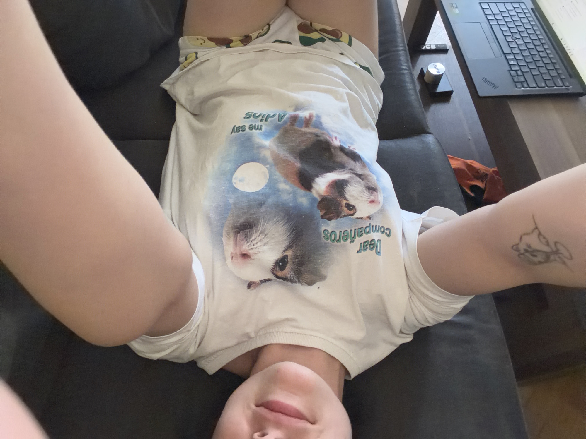A person is lying on their back on a dark couch, with their head near the bottom of the frame and their feet toward the top. They are wearing a white t-shirt with a graphic of guinea pigs and text that reads "Dear compañeros me say Adios" printed on it. The t-shirt appears to be slightly stretched around the shoulders. The person has light skin and their face is partially visible at the bottom of the image, with their eyes closed and a slight smile. Their arms are extended upwards, with their left arm resting on their chest and their right arm extended to the right side. A tattoo of a cartoon cat is visible on their right upper arm.
To the right of the couch, a dark wooden table is visible. On the table, there is a black laptop with a keyboard, a small cylindrical object with writing on it, and a black rectangular object. An orange piece of fabric is draped over the side of the table. The environment appears to be indoors, with natural light coming from the right side of the frame. The overall scene suggests a casual, relaxed setting.