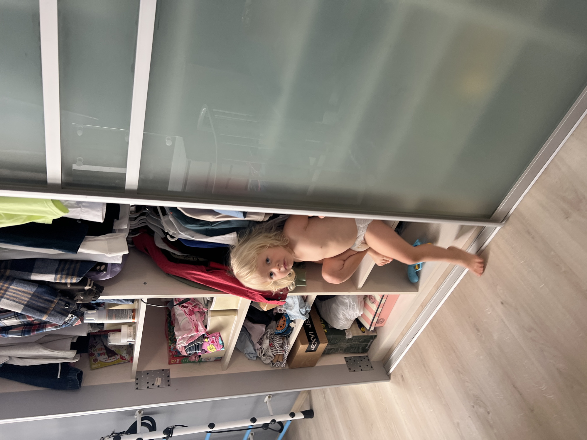 A young child with blonde hair and fair skin is lying down inside an open closet. The child is wearing a diaper and appears to be looking at the camera with a curious expression. The closet is filled with clothes and other items, suggesting it is a storage space within a home. The scene is well-lit, possibly indicating daytime. The child's relaxed posture and the presence of toys scattered on the shelves suggest a casual, at-home environment. The image is taken from a slightly elevated angle, looking down into the closet. The closet doors, made of frosted glass, are visible at the top of the frame. The flooring visible outside the closet is light-colored wood.