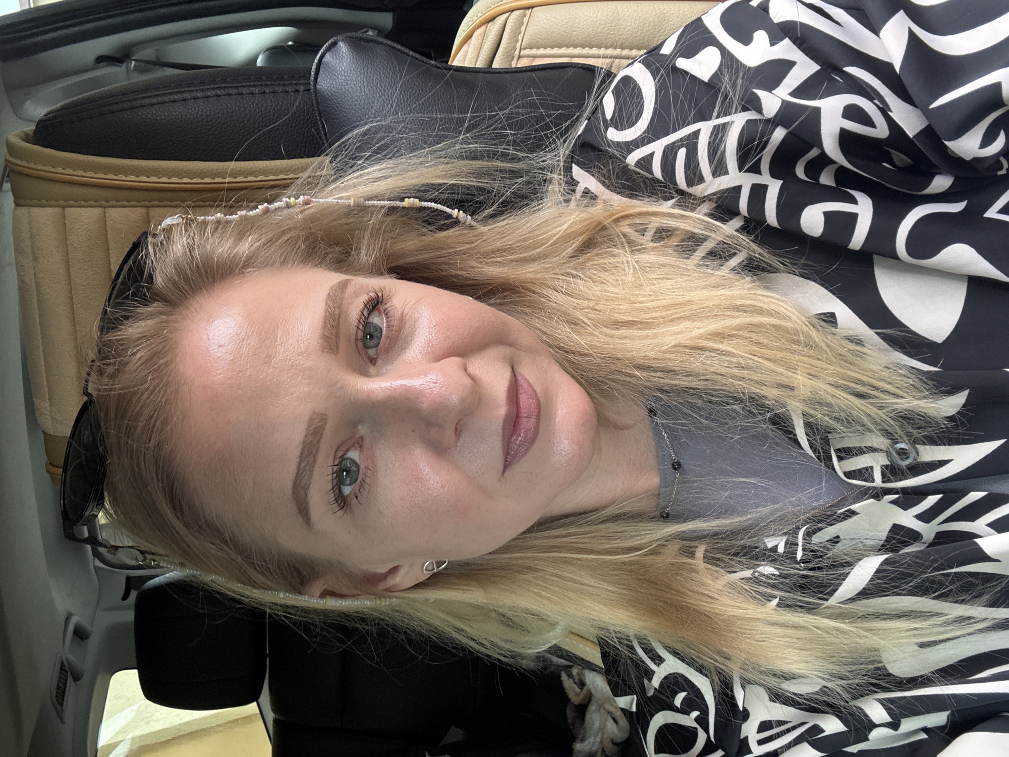 The image is a close-up portrait of a light-skinned woman with blonde hair, likely inside a vehicle. She is smiling and looking at the camera. She is wearing a black and white patterned garment. A string of beads adorns her hair near the forehead. Part of a car seat is visible in the background. Sunglasses are also visible near her head. The image appears to be taken in Unknown, Unknown.