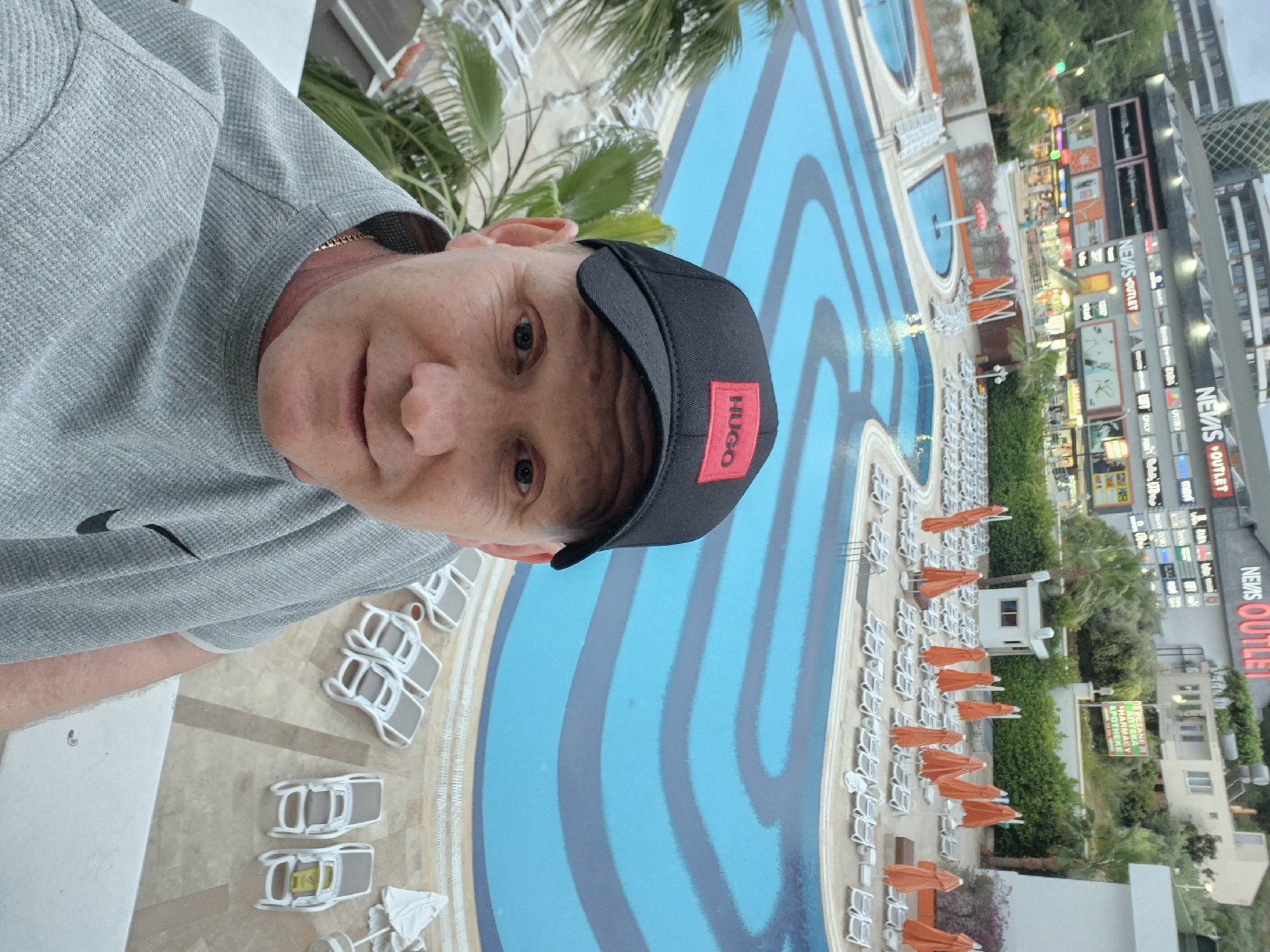 A selfie of a man wearing a grey shirt and a black cap with the word "HUGO" in red, taken from an elevated position. In the background, there is a large swimming pool with a striped blue and grey pattern. Rows of white beach chairs, some with orange umbrellas, are arranged alongside the pool. In the distance, there is a multi-story building with signs indicating commercial establishments, including one that reads "NEWS OUTLET". The scene appears to be a tourist resort in Alanya, Turkey, based on the architectural style, vegetation, and commercial signs.