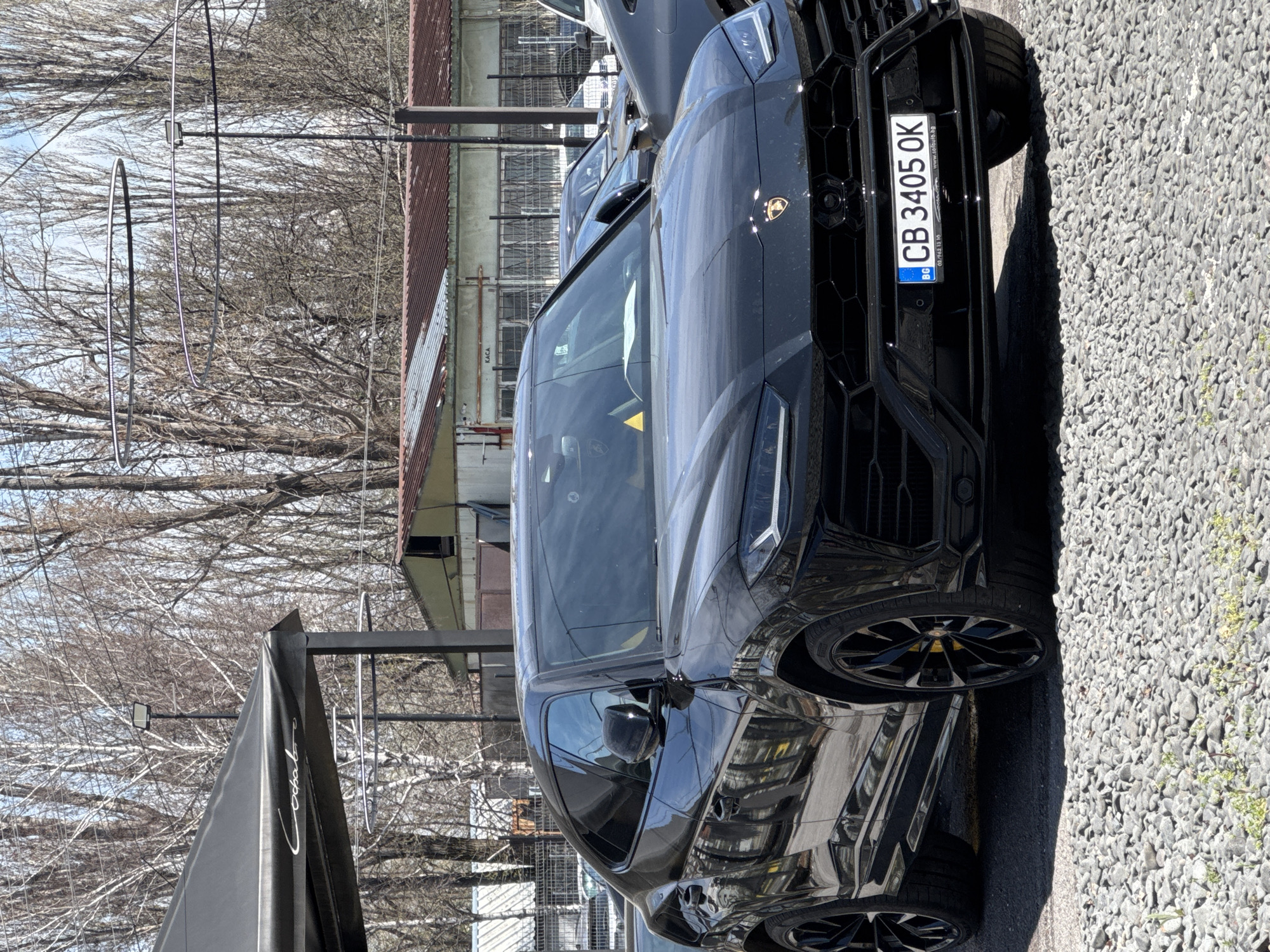 The image depicts a black Lamborghini Urus parked on a gravel lot. The license plate reads "CB 3405 OK" and indicates it is registered in Bulgaria. The background includes bare trees, buildings, and overhead wires. Based on the license plate and general architectural style, the location is likely within the city of Sofia, Bulgaria. No people are clearly visible in the image.