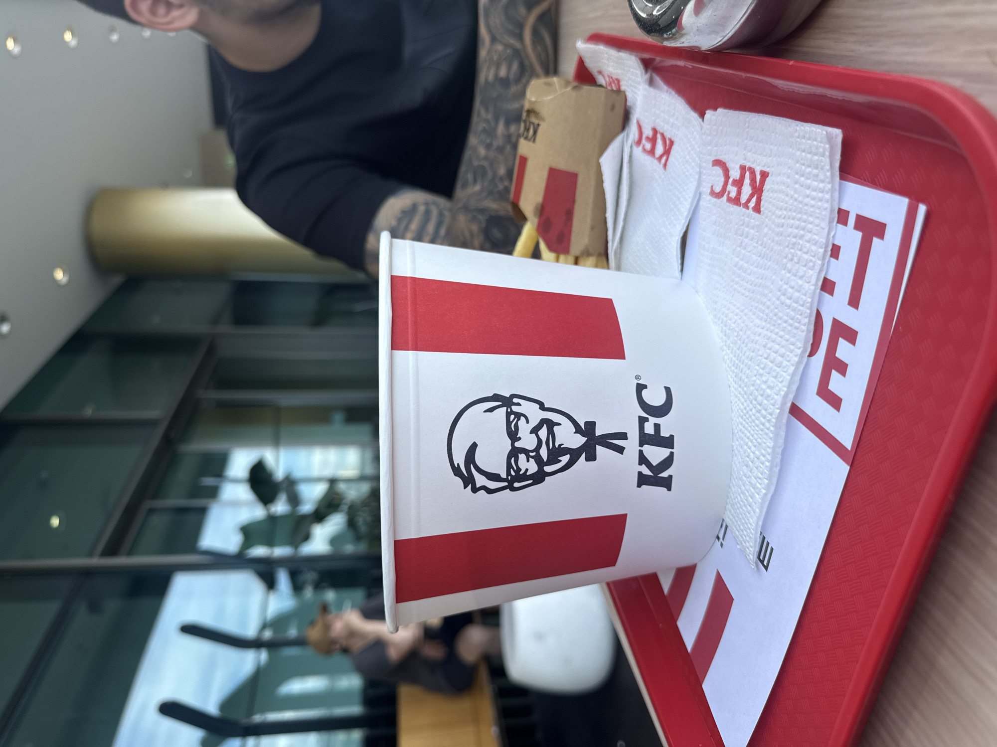 In the foreground, a red plastic tray holds a white KFC bucket with the brand's iconic Colonel Sanders logo and "KFC" text. French fries are partially visible next to a small brown KFC-branded paper container. Several white KFC-branded napkins are layered on the tray. Behind the KFC bucket, the blurred figure of a person with tattooed arms wearing a black t-shirt is seated at a table. In the background, a person wearing a cap is seated at a table near a large window with reflections of trees and sky. The setting appears to be an indoor dining area with modern architecture, possibly a KFC restaurant in Burgas, Bulgaria, judging by the stylistic elements of the building's interior and exterior, such as the large glass windows, polished floors, and modern column.
