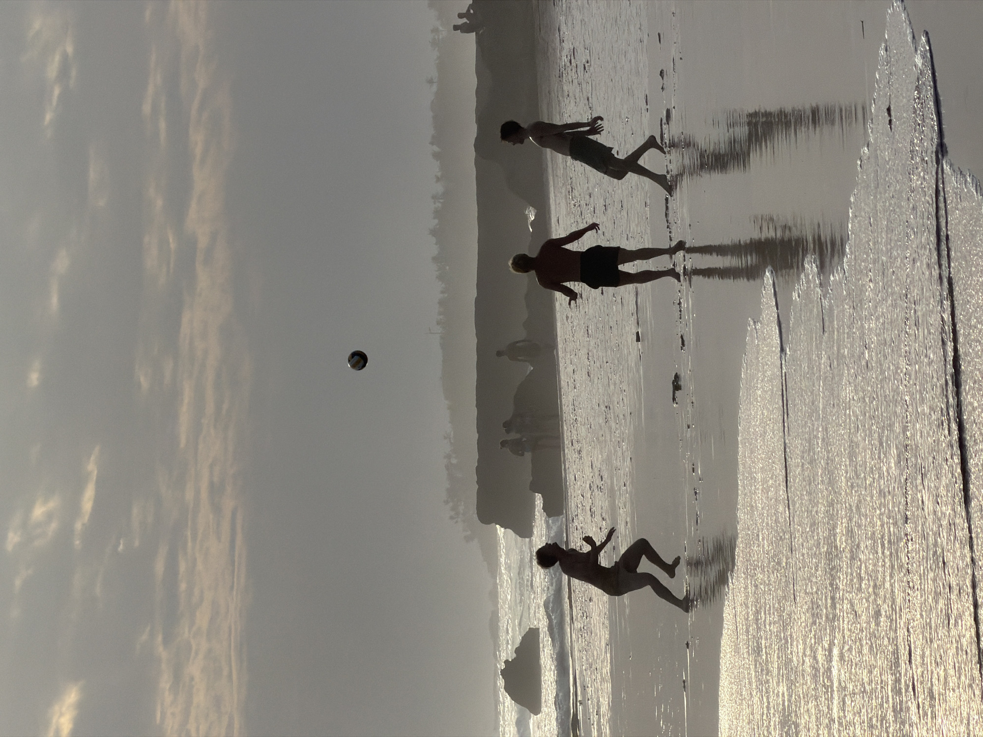 The image captures a group of young people playing volleyball on a beach at sunset, likely in North Kuta, Indonesia. Silhouetted against a hazy sky with scattered clouds, three individuals are prominently featured in the foreground, actively engaged in the game. One person, positioned on the left, is in mid-air, appearing to serve or strike the ball. Another individual, in the center, is facing away from the viewer, also in motion, as if ready to receive the ball. The third person, on the right, is looking upwards, tracking the ball's trajectory. A volleyball is visible in the air above them.
The beach is wet and reflective, with gentle waves lapping at the shore, creating shimmering patterns of light. In the mid-ground, several other silhouetted figures are visible near rocky outcrops and vegetation that line the coastline, suggesting a popular beach location. The background is dominated by a soft, diffused light characteristic of a sunset or sunrise, with a distant, hazy landmass forming the horizon. The overall atmosphere is serene and energetic, highlighting a moment of recreation amidst a natural coastal setting.