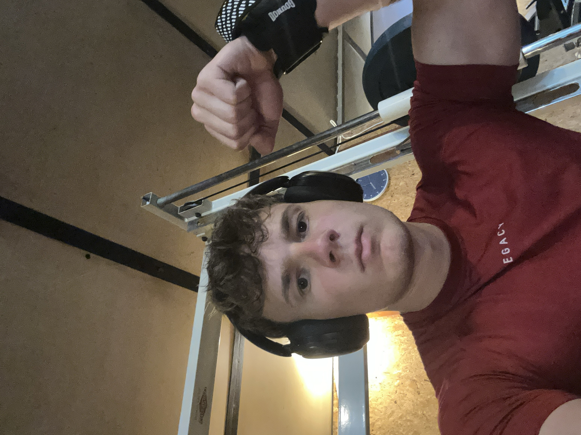 A man wearing a red shirt and headphones is in a gym. He is next to a weight machine with weights loaded on it. He is wearing a wrist wrap on his left hand. The image appears to be taken from a low angle. The location is not evident from the image.
