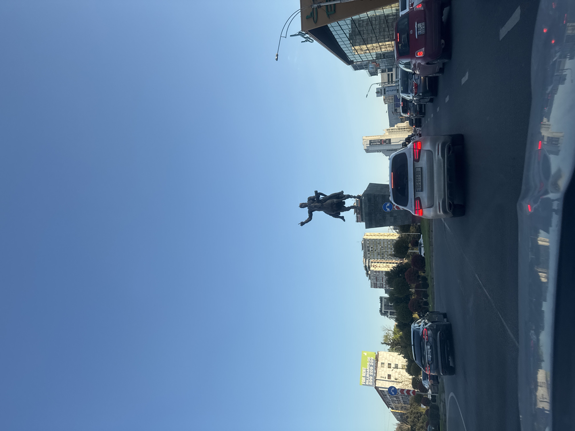 The image displays an urban street scene in Tbilisi, Georgia, under a clear blue sky. The original image is rotated ninety degrees clockwise; when viewed correctly, it shows a multi-lane road with an equestrian statue as a central feature.
In the mid-ground, a large bronze equestrian statue of a historical figure stands on a stone pedestal. The statue depicts a man on horseback, with one arm outstretched. On the road, a line of cars is visible, suggesting traffic or a stop. In the foreground, the rear of a silver SUV with the Georgian license plate "BG-017-BB" is prominent. Further down the road, a red Fiat 500 with a partially visible Georgian license plate "GE-909-RT" is also in traffic, followed by other vehicles.
The background consists of city architecture, including modern buildings with reflective glass facades and older, multi-story residential blocks. Green trees and bushes line the road in some areas. The weather appears sunny and clear, with bright daylight and visible shadows, indicating either morning or afternoon.
Notable details include:
- **Visible Text:** The license plates "BG-017-BB" and "GE-909-RT" are visible on the vehicles. A large billboard on the right side of the road reads "FOR RENT BILLBOARDS.GE 599 209 209".
- **Road Signs:** Several blue circular road signs with white arrows indicating mandatory directions are present.
- **Location Cues:** The Georgian license plates and the ".GE" domain on the billboard confirm the location as Tbilisi, Georgia.
No individual people are distinctly visible, only the historical statue and vehicles in traffic.