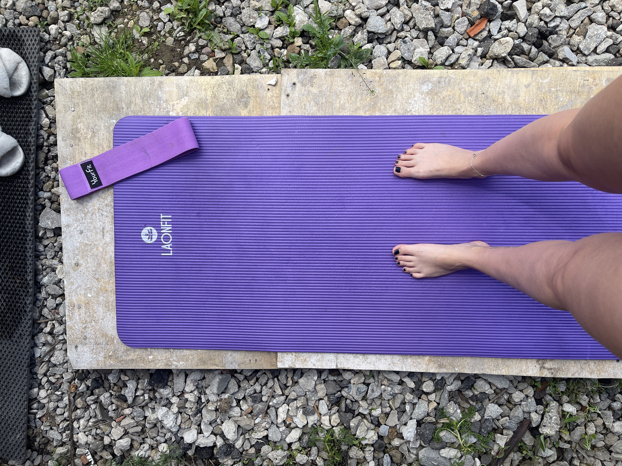 The image shows a purple yoga mat placed on a rough concrete surface amidst small grey rocks and some scattered greenery. A purple resistance band is situated near the top left corner of the mat. A person stands on the mat, their feet visible in the center of the image, with black nail polish. The location seems to be an outdoor space in Мосметаллоконструкция Онт, Russia. To the left of the concrete surface is a dark-colored, textured pad or mat with white fuzzy objects possibly slippers.