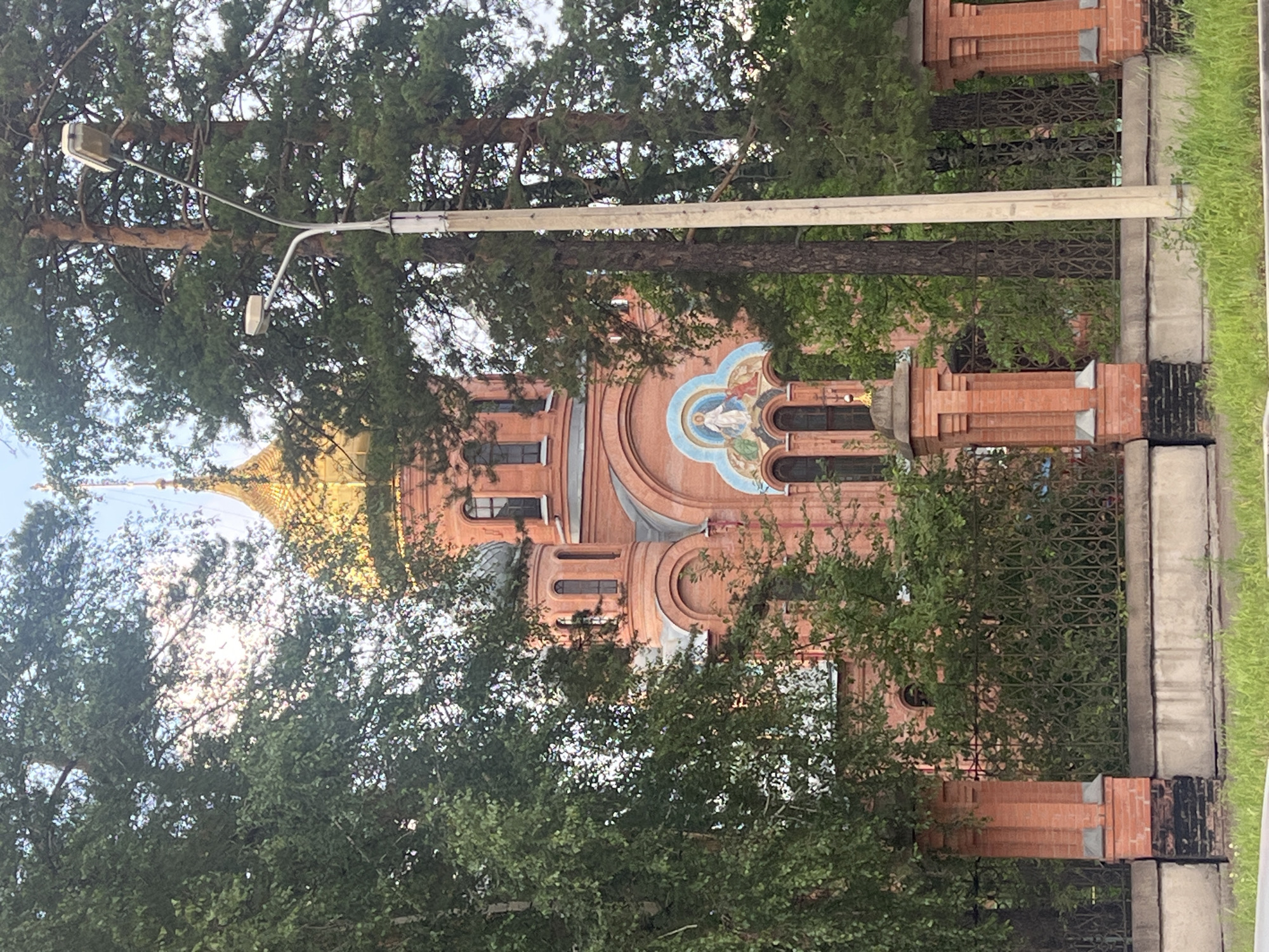 The image shows a brick church with a golden dome, partially obscured by trees. The church has arched windows and a mosaic above an entrance. A metal fence and brick pillars are in the foreground. A street light is visible on the right side of the image. The scene is located in Zheleznogorsk, Russia.