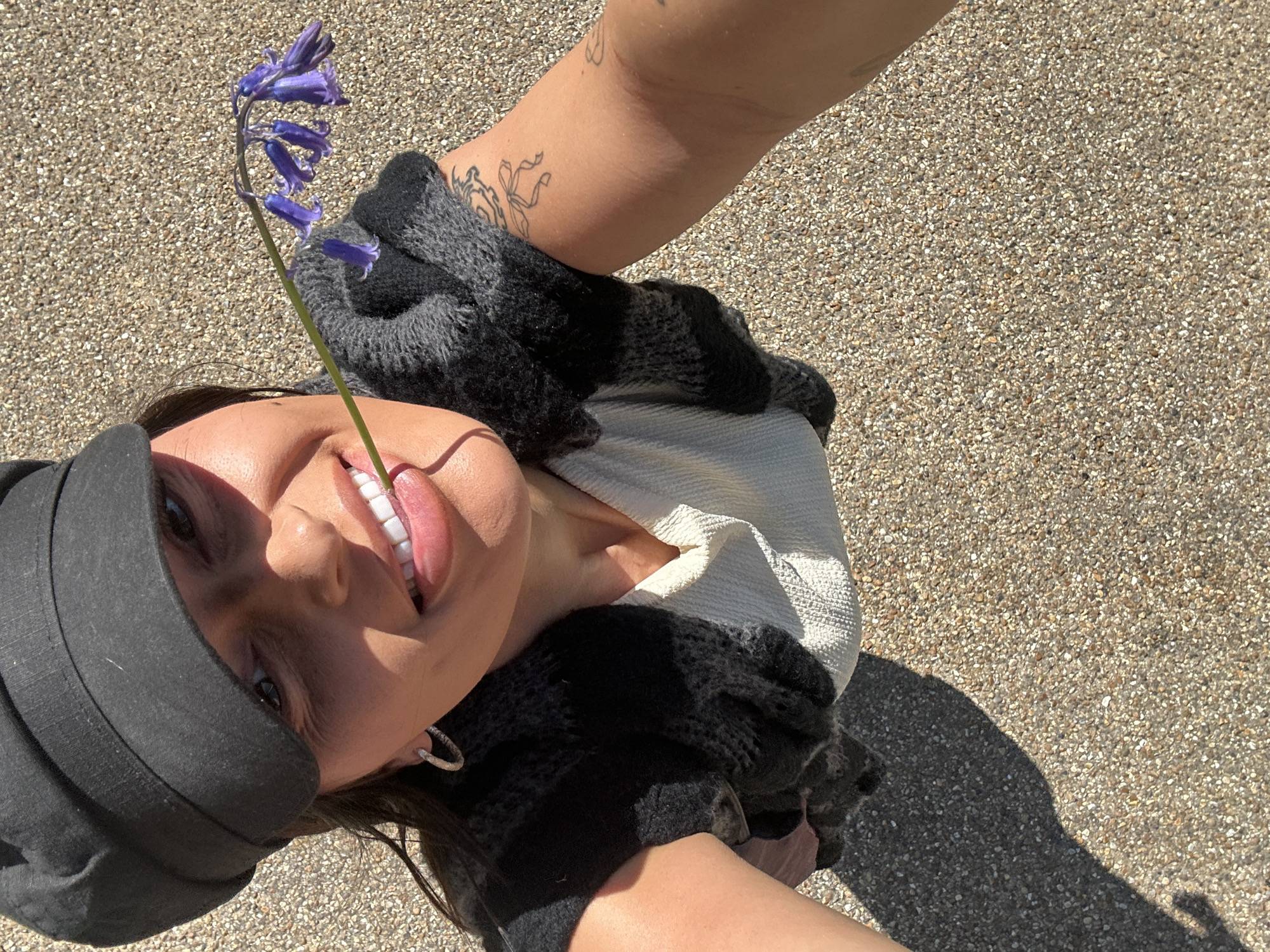 A light-skinned female wearing a black cap and gray knit gloves is holding a bluebell flower in her mouth. She has an earring on her left ear, and a tattoo on her right forearm. She's holding the flower between her teeth and sticking her tongue out. The ground is light brown gravel. The image appears to have been taken in daylight. The location is Tunbridge Wells, United Kingdom.