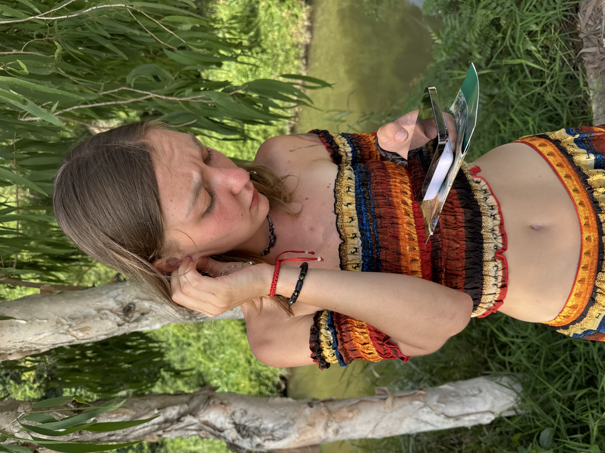 A young woman stands outdoors, in a natural setting with trees and a body of water. She is wearing a colorful, off-the-shoulder top and a matching skirt. Her hair is brown and pulled back. She is looking down at her phone, which she is holding in her right hand. In her left hand, she is touching her ear. She has several bracelets on her wrists and a necklace around her neck. The environment suggests a location with lush vegetation and a tranquil atmosphere. The specific location, Nam Hoa Lư Ward, Vietnam, indicates a potential connection to the historical and cultural significance of the area, possibly referencing the Hoa Lư ancient capital.
