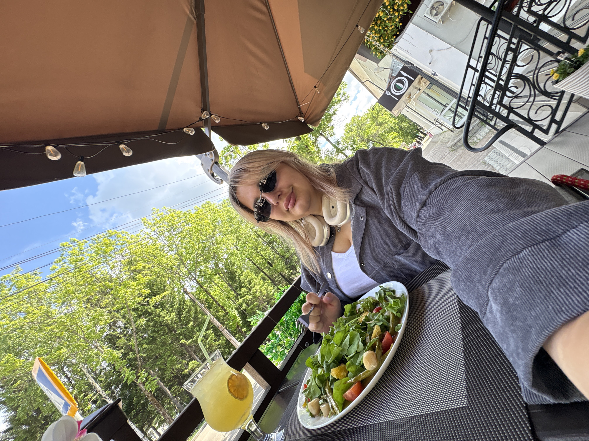 In an outdoor seating area, a young woman with blonde hair and sunglasses is taking a selfie with her meal. She is wearing a grey jacket over a white top and beige headphones around her neck. In front of her is a white oval plate filled with a green salad containing cherry tomatoes, what appear to be white cheese cubes, and green beans. To the left of the salad, a tall glass of yellow liquid, likely lemonade, with a slice of citrus fruit and a straw rests on the table. The table is covered with a dark grey and white patterned placemat. Behind the table, trees with lush green foliage are visible against a bright blue sky with scattered clouds. A brown awning with string lights is visible overhead, suggesting a restaurant or cafe setting. The background includes a building with a sign depicting a fork and knife, indicating a dining establishment. This scene is likely located in Birobidzhan, Russia, given the context.