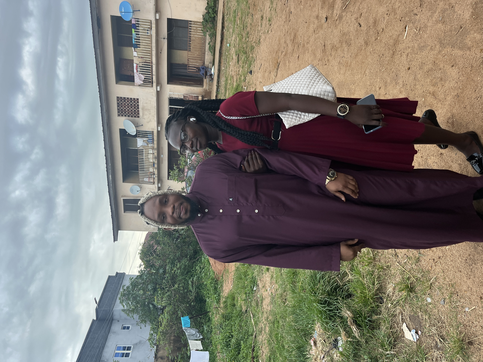 In Jos, Nigeria, a man and a woman stand facing forward in an outdoor setting with a building in the background. The man, wearing a maroon robe, has blonde braided hair and a beard. The woman, in a dark red dress, wears glasses, has dark braided hair, and carries a white quilted purse. They are standing on a dirt path surrounded by grass and some debris. The building behind them is a two-story structure with balconies and satellite dishes. The sky is overcast.
