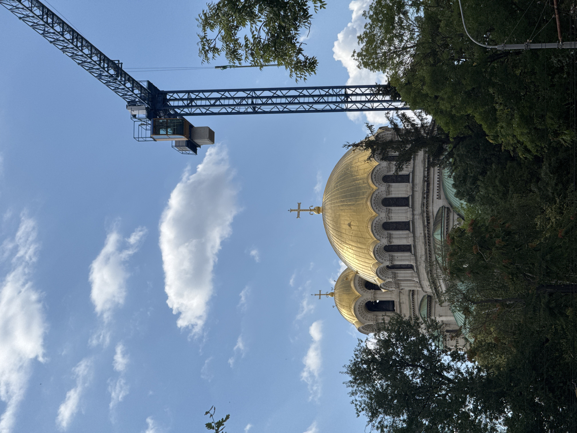 The image shows the Alexander Nevsky Cathedral in Sofia, Bulgaria, under a blue sky with white clouds. A large construction crane is positioned to the right of the cathedral, with its arm extending over the building. The golden domes of the cathedral are visible, as well as trees and some parts of the surrounding urban environment, including a lamppost. The scene suggests ongoing construction or renovation work near the historical landmark.
