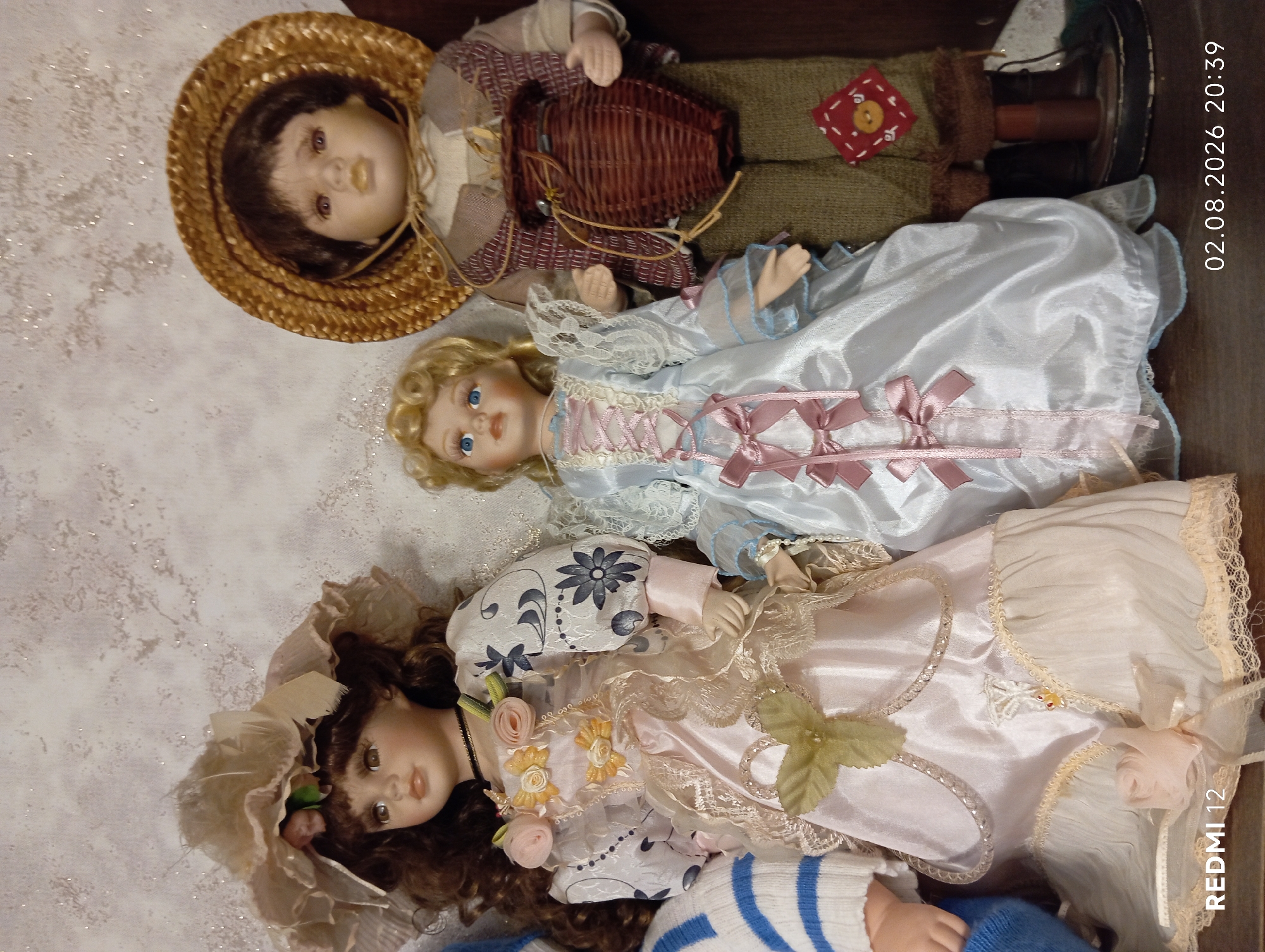 Three porcelain dolls are displayed on a shelf against a textured, off-white wall. The doll on the left wears a light pink satin dress with lace trim and a wide-brimmed hat adorned with fabric flowers. The central doll is dressed in a pale blue satin dress with pink ribbon accents. The doll on the right, appearing to be a boy, wears a straw hat, a vest over a cream-colored shirt, and green pants with a red diamond patch on the right leg. He holds a small woven basket. The shelf appears to be made of wood. The image is watermarked with "REDMI 12" and "02.08.2026 20:39". The location of Oryol, Russia is not depicted in this image.