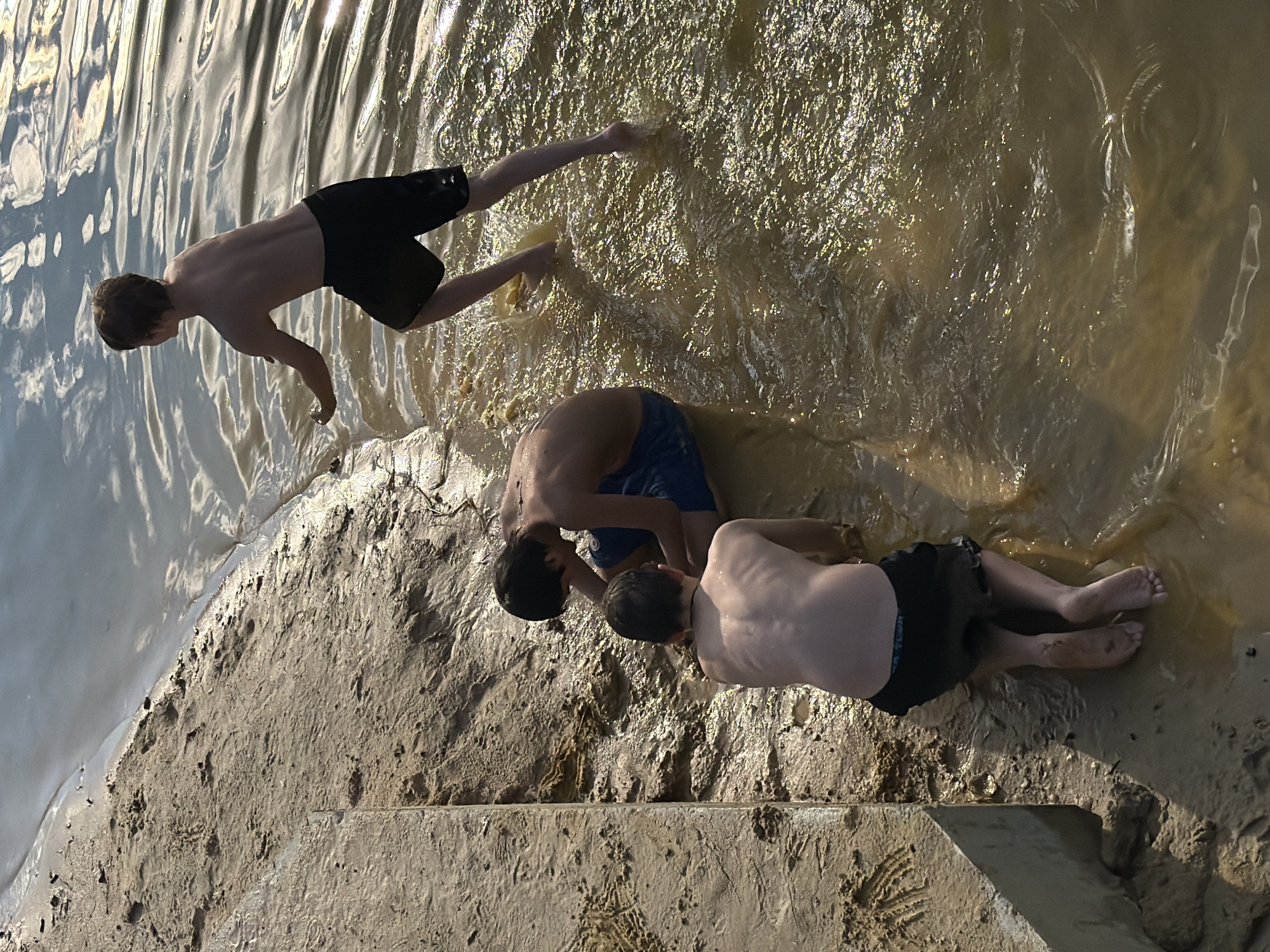 Three young males are interacting near a body of water, likely a river or lake, in Udelnaya, Russia. The terrain is a mix of sandy, muddy ground and shallow water. The water is murky and reflects the sunlight, creating shimmering patterns. One boy, on the right, stands in the water, facing away from the camera, with his arm outstretched as if throwing something. Another boy is crouched in the water, looking down at the ground. The third boy is kneeling on the wet sand, facing away from the camera and towards the water. All three are shirtless, wearing shorts. A concrete structure runs along the left edge of the sandy bank. The overall scene depicts a casual, outdoor activity in a natural setting.