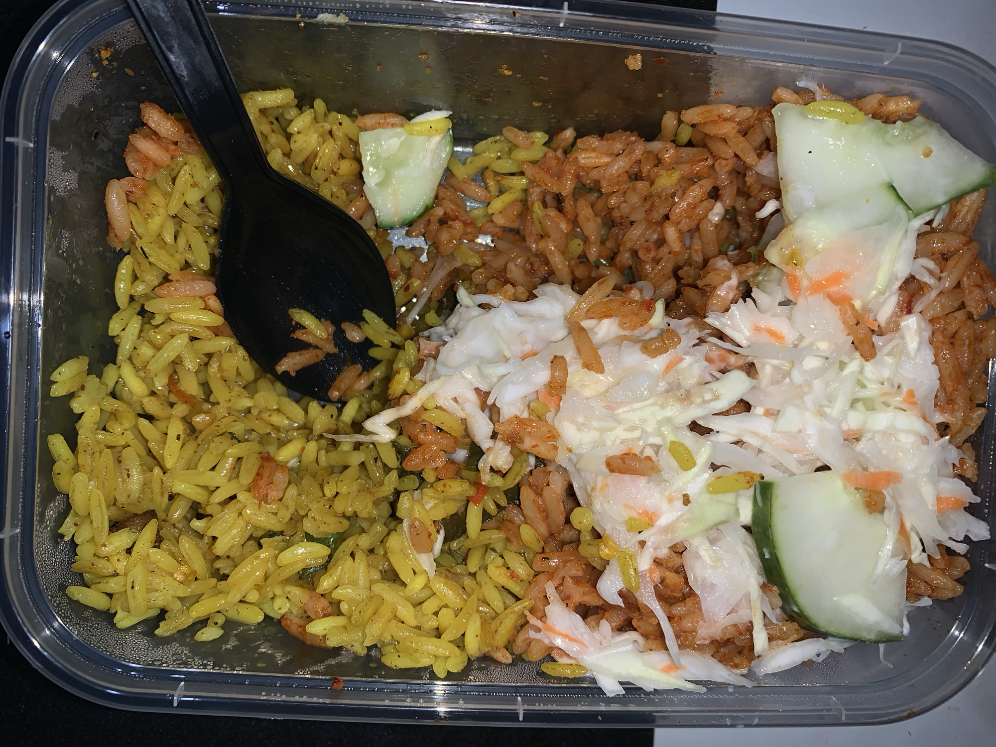 An overhead close-up shot captures a meal packed in a clear plastic takeout container, likely in Rafin Par, Nigeria. The container is filled with a vibrant assortment of food, neatly partitioned.
On the left side, a substantial portion of bright yellow-tinted rice, possibly turmeric rice or a seasoned rice, is visible. Small reddish-orange pieces are interspersed throughout the yellow grains, suggesting spices or tiny protein elements. A black plastic spoon is partially inserted into this section of rice.
The right side of the container holds a generous amount of reddish-orange rice, consistent with the appearance of jollof rice. Layered over both rice types and centrally placed is a fresh, crisp slaw composed of shredded white cabbage and orange carrot slivers. Several thick, green-skinned cucumber slices are arranged on top of the slaw and rice, providing a cooling garnish.
The clear plastic container indicates the meal is prepared for takeaway or casual dining. The food appears ready for consumption. There are no people or discernible text visible in the image.