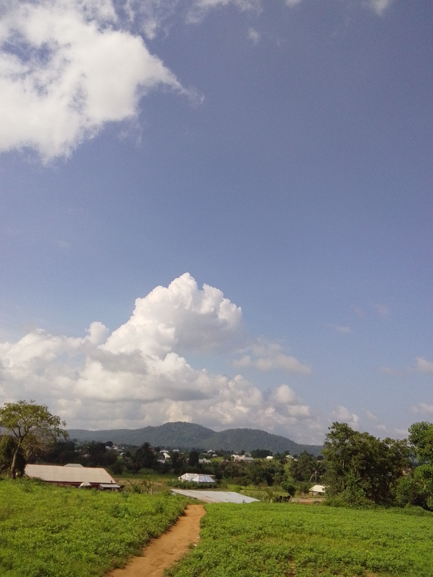The image depicts a rural landscape with a clear blue sky and scattered white clouds. In the foreground, a dirt path winds through a lush green field. To the left of the path, there is a long, low-lying building with a light-colored roof. In the middle ground, several other buildings are visible, interspersed with trees and vegetation. A range of hills forms the backdrop of the scene, and a small village appears to be nestled at the foot of the hills. The overall impression is one of tranquility and natural beauty, consistent with a location in the city of Serti, Nigeria, where such scenic vistas are common.