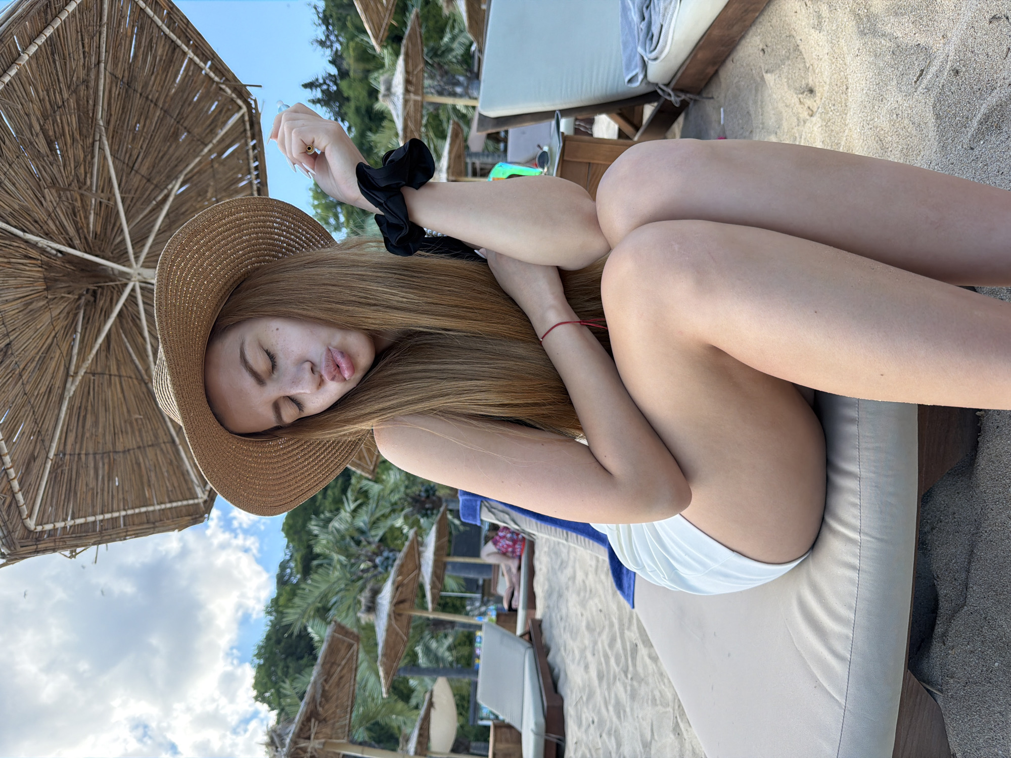 This is a vertical, close-up, eye-level shot of a young woman lying on a beach lounge chair. She is wearing a straw hat, a black top, and white bikini bottoms. Her eyes are closed, and her lips are pursed as if she is blowing a kiss. Her long, blonde hair spills out from under the hat, and a black scrunchie is visible on her arm. A red string bracelet is tied around her wrist. The background shows a beach with sand, palm trees, thatched umbrellas, and other lounge chairs. The sky is blue with some clouds. The lighting suggests it is daytime. The overall mood is relaxed and playful.