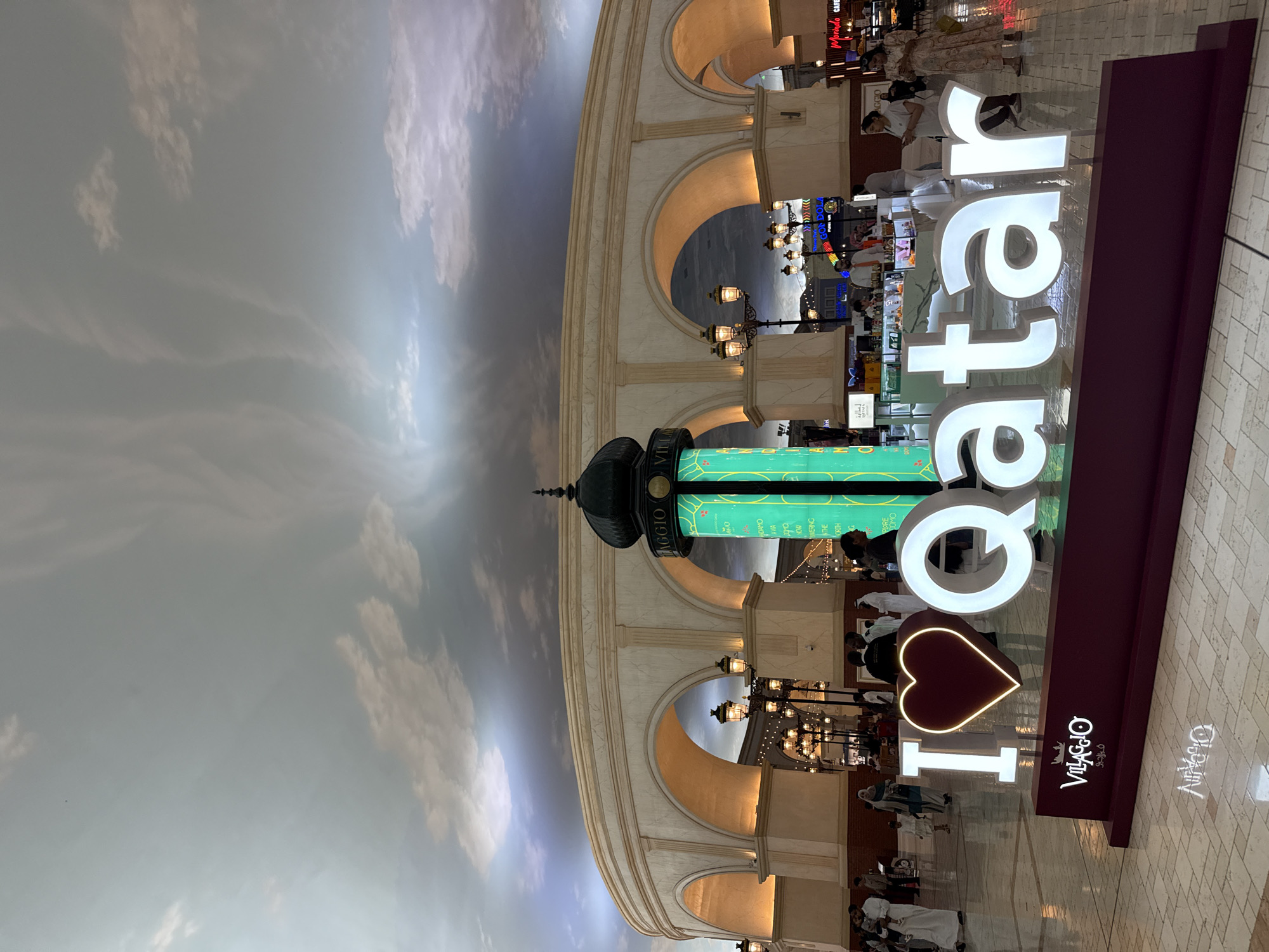 The image shows the “I love Qatar” sign at the Villaggio Mall in Al Rayyan, Qatar. The foreground includes the sign, constructed with a heart shape replacing the word "love", and “Qatar” stacked vertically in large, illuminated white letters. The sign's base is a dark red or maroon color and features the words "Villaggio" and "فيراجيو" (Villaggio in Arabic).
In the background, the architectural style of the Villaggio Mall is visible, featuring arches and facades that mimic Venetian architecture. There are decorative lamp posts and glimpses of shops and restaurants lining the mall’s walkways. Several people are seen walking, shopping, or sitting at outdoor cafes.
The ceiling of the mall is painted to resemble a cloudy sky. The floor is tiled in a light color. The overall lighting suggests a bright, indoor environment designed to mimic an outdoor setting.