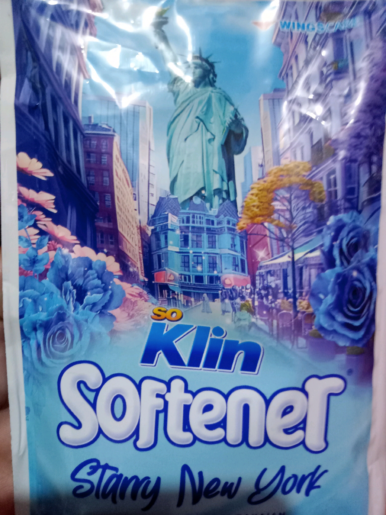 The image is a close-up shot of a product package, likely for fabric softener, featuring a stylized illustration of New York City. The illustration depicts the Statue of Liberty in the background, surrounded by tall buildings and street-level scenes with people and cafes. Prominent blue and pink flowers are integrated into the design, creating a dreamy and somewhat abstract aesthetic. The product's branding includes the words "So Klin" and "Softener," with "Starry New York" as a descriptive subtitle. The packaging itself is predominantly light blue with white text and illustrations. The photo is taken from a slightly elevated angle, capturing the entire package which is illuminated by overhead lighting, causing some glare and reflections on the plastic surface. There is no visible indication of any specific location in East Jakarta, Indonesia, as the image focuses entirely on the product design.