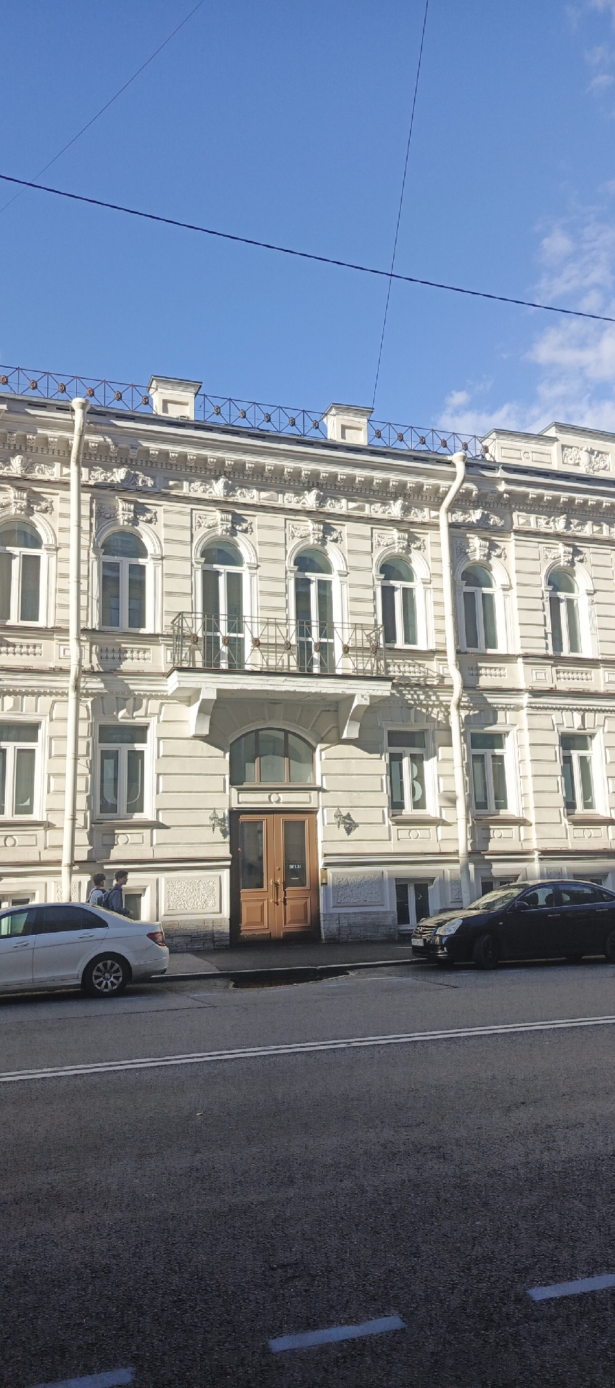 A low-angle, eye-level shot shows the ornate facade of a light-colored building in Saint Petersburg, Russia. The building exhibits classical architectural elements, including arched windows with decorative molding and a detailed cornice. A wrought-iron railing runs along the roofline. A white drainpipe runs vertically down the left side of the facade. Below the building, a street runs parallel to it, with two cars parked on the right side and one car parked on the left. Two young men in casual clothing are walking on the sidewalk in front of the building's entrance. The sky above is a clear, bright blue with a few wispy clouds and is crossed by two dark wires. The image captures a typical street scene in a historical district of Saint Petersburg.