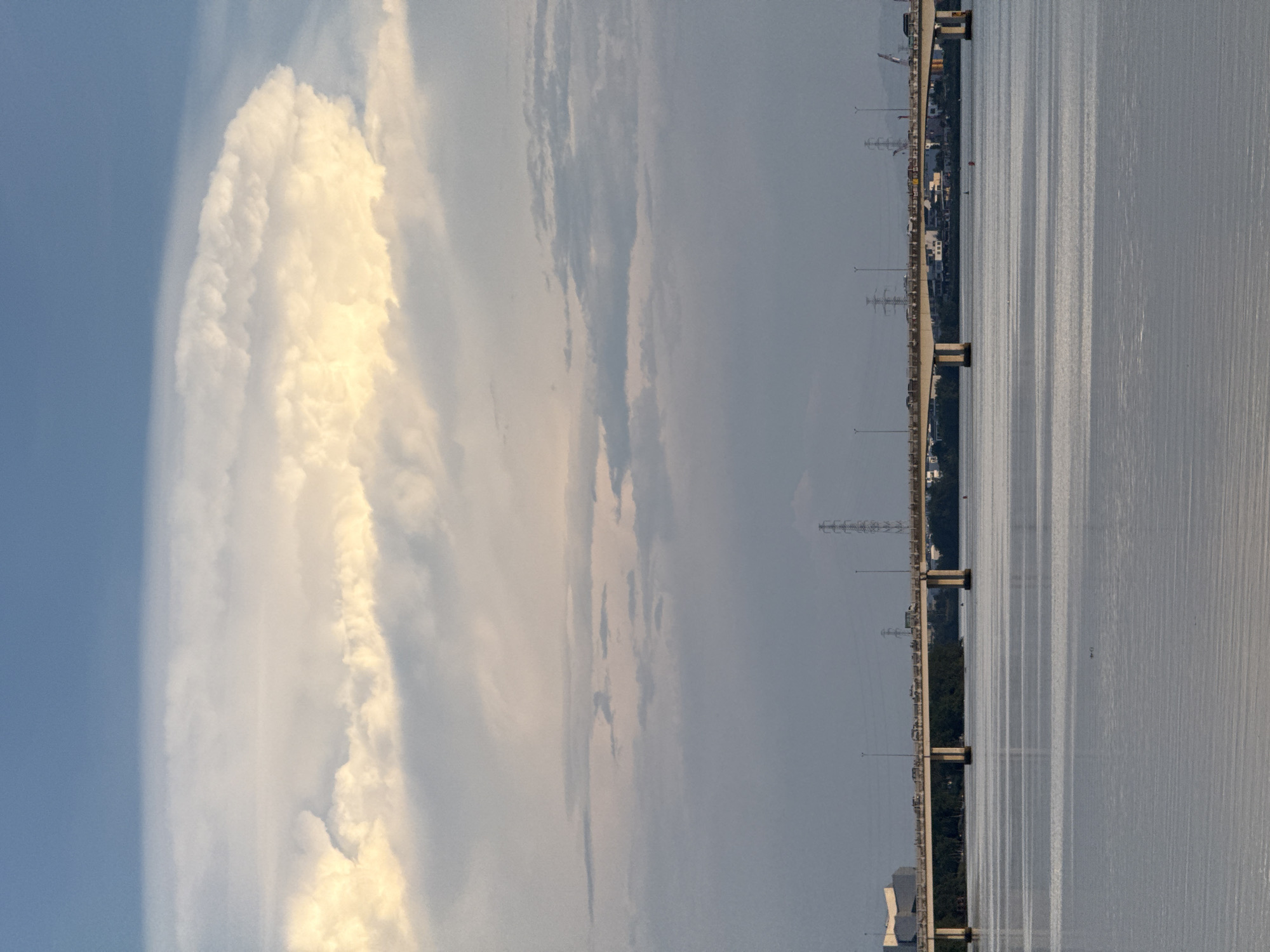 The image captures a wide shot of the Han River in Đà Nẵng, Vietnam, under a dramatic sky. A long, concrete bridge, likely the Han River Bridge, spans the width of the frame, carrying a heavy flow of traffic. The water in the foreground is calm, reflecting the overcast sky. In the distance, the cityscape is visible, dotted with buildings and power lines, hinting at urban development. The sky is a prominent feature, with a large, luminous cloud formation dominating the upper portion of the image, tinged with warm light, contrasting with the softer, grayer clouds below. The overall atmosphere is serene yet suggests the bustle of a vibrant city.