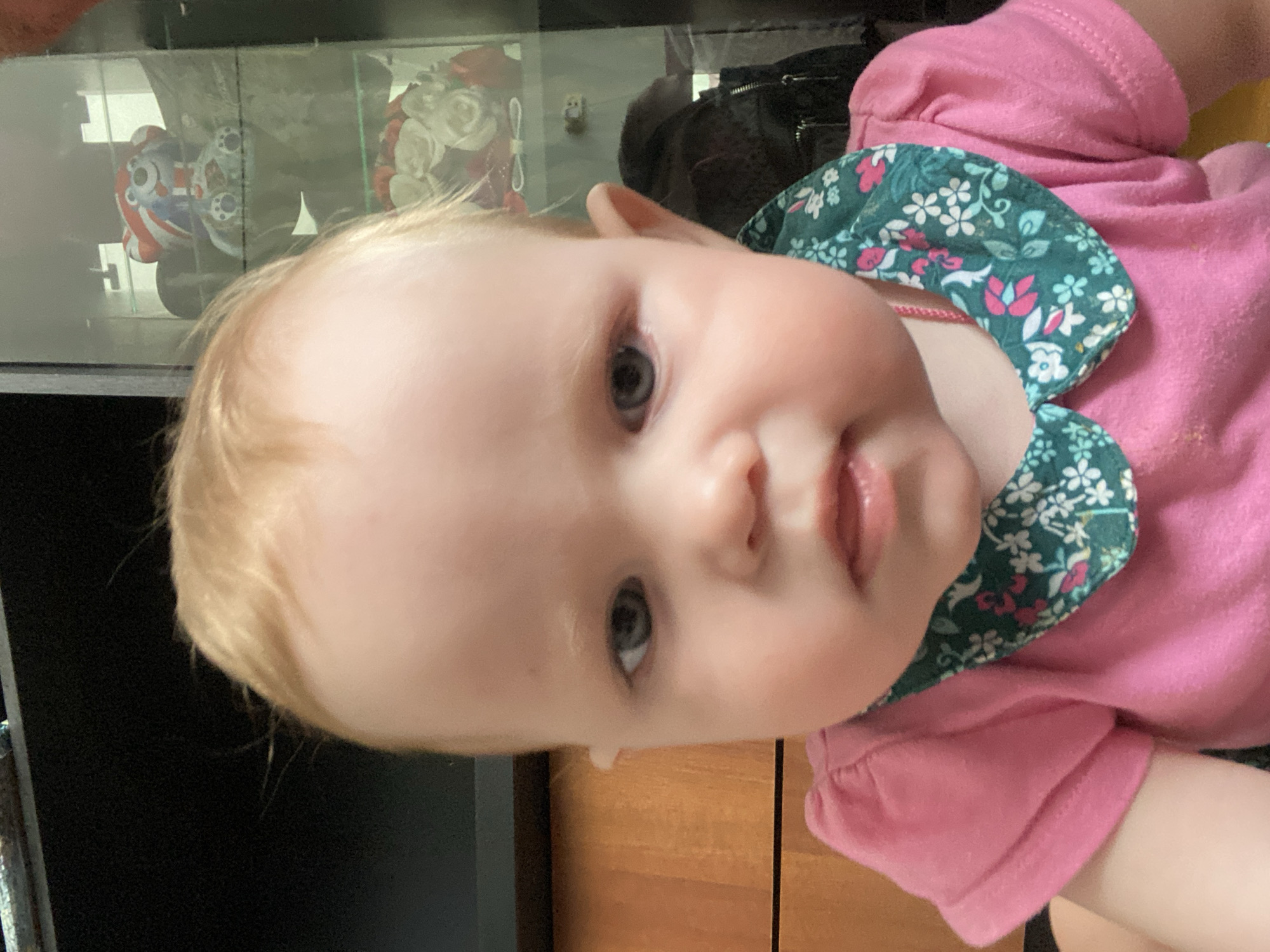 A close-up shot captures a fair-skinned baby with light blonde hair and grey-blue eyes. The baby wears a pink shirt with a dark green collar patterned with white and red flowers. The baby's face is slightly blurred. Behind the baby, there's a dark object and a glass display case containing a ceramic figure with red, white, and blue colors, alongside a floral arrangement in the display case. The wooden surface is visible in the background. The location in the city of Фоки, Russia is not possible to confirm from the image provided.