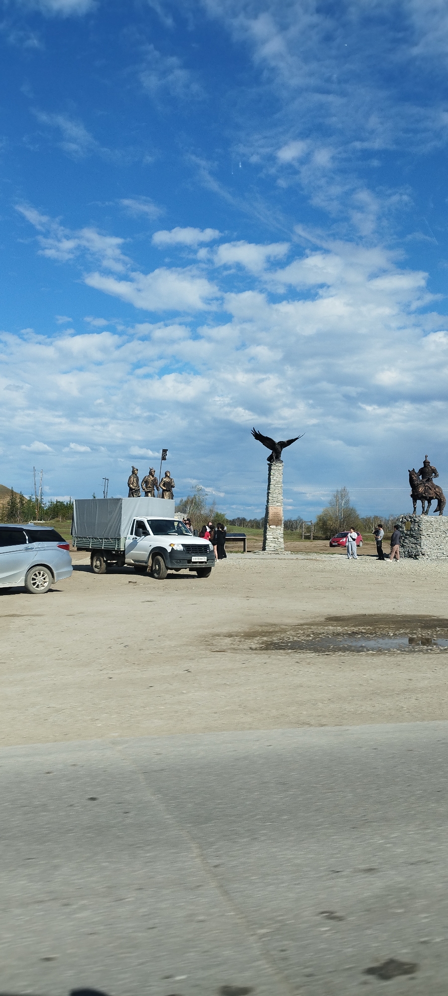 The image depicts a scene in the city of Tekhtyur, Russia. In the foreground, there is a section of paved road. The main focus is a sandy, unpaved area where several items of interest are located. A silver SUV is parked on the left side of the frame. Next to it is a white UAZ truck with a gray canvas covering the truck bed. Three statues of warriors, possibly bronze, stand atop the covered truck bed. To the right, two monuments are visible. One features a large, black eagle perched on top of a stone column. The second monument portrays a warrior on horseback. Several people are present in the area, appearing to be tourists or visitors. Some are standing near the monuments, while others are close to the truck. The background includes trees and distant hills. The sky is bright blue with scattered white clouds.