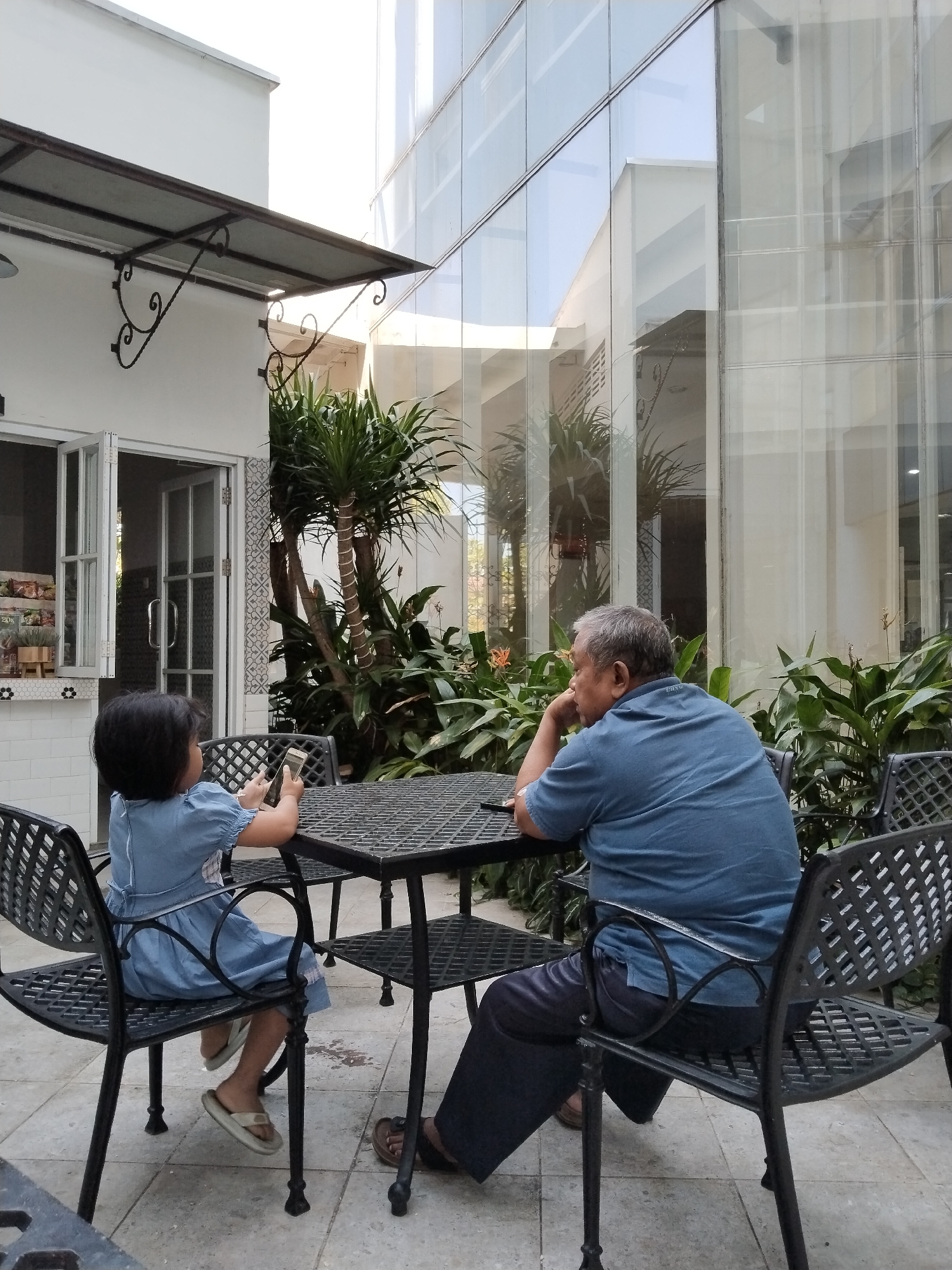 An older man in a blue short-sleeved shirt and dark pants sits at a metal table outdoors, facing away from the camera, with his elbow on the table and his hand on his chin. Across from him, a young girl in a blue dress sits on a matching metal chair, holding a smartphone. The table and chairs are on a tiled patio in front of a building with a white facade and large glass windows. Lush green plants are visible behind the table and chairs. In the background, a modern building with glass panels reflects the sky. The scene appears to be a cafe or restaurant patio in Kecamatan Pati, Indonesia, during daylight hours.