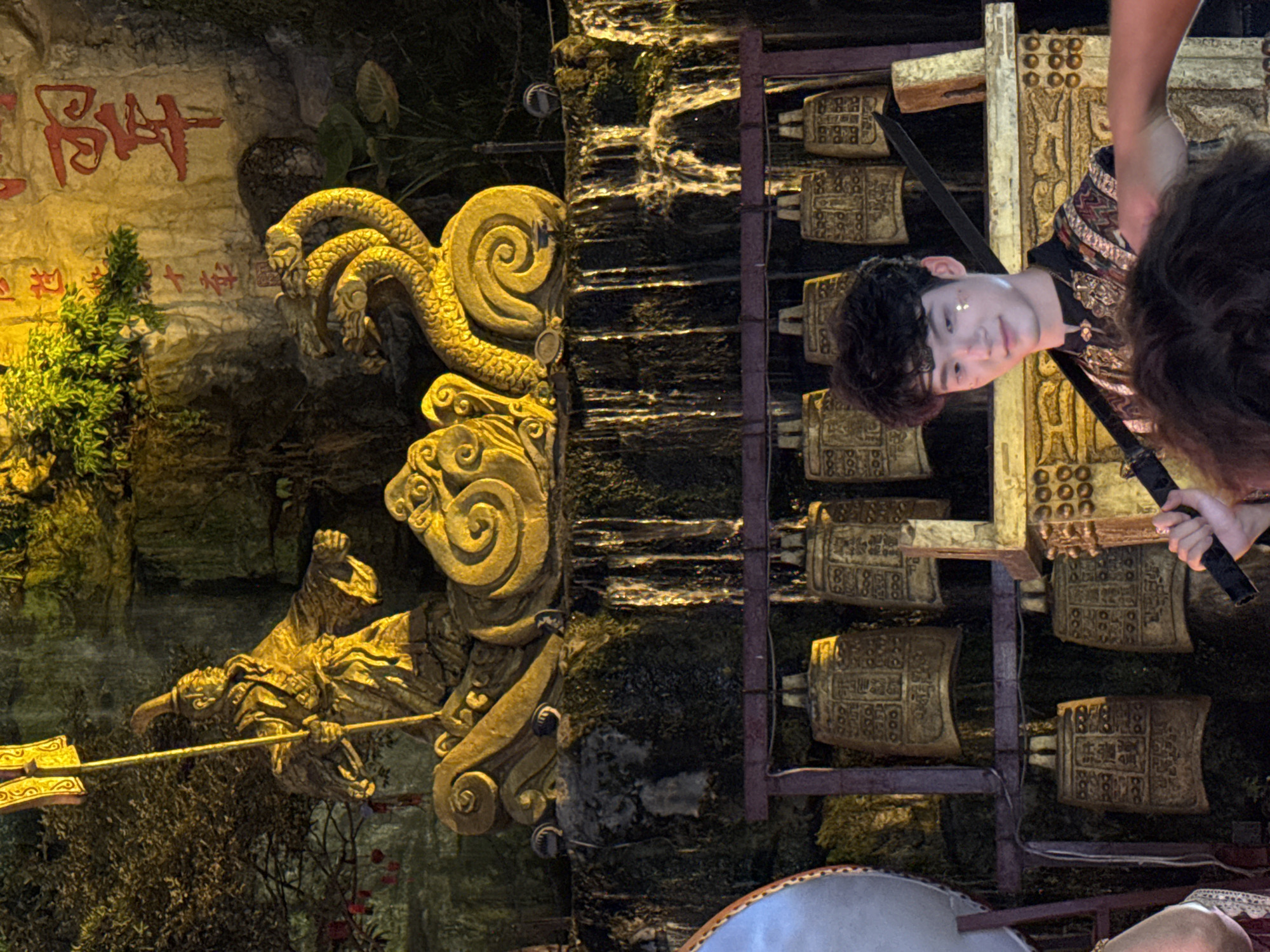 The image captures a performance setting in Yuzhong District, China, likely at a cultural or historical site. In the upper portion, a golden statue of a robed figure holding a flag stands atop a decorative base with swirling motifs and a three-headed dragon sculpture. Behind the statue, a rock face features Chinese calligraphy in red. A man with dark, curly hair, adorned with glitter on his face and wearing a black and gold patterned garment, stands in the foreground holding a black sword. He is looking towards the viewer. Behind him, a row of ancient-looking bronze bells hangs on a rack. Water is cascading down the rock face behind the statue. The lighting is dramatic, with warm yellow and green hues illuminating the scene, suggesting an evening or indoor performance.