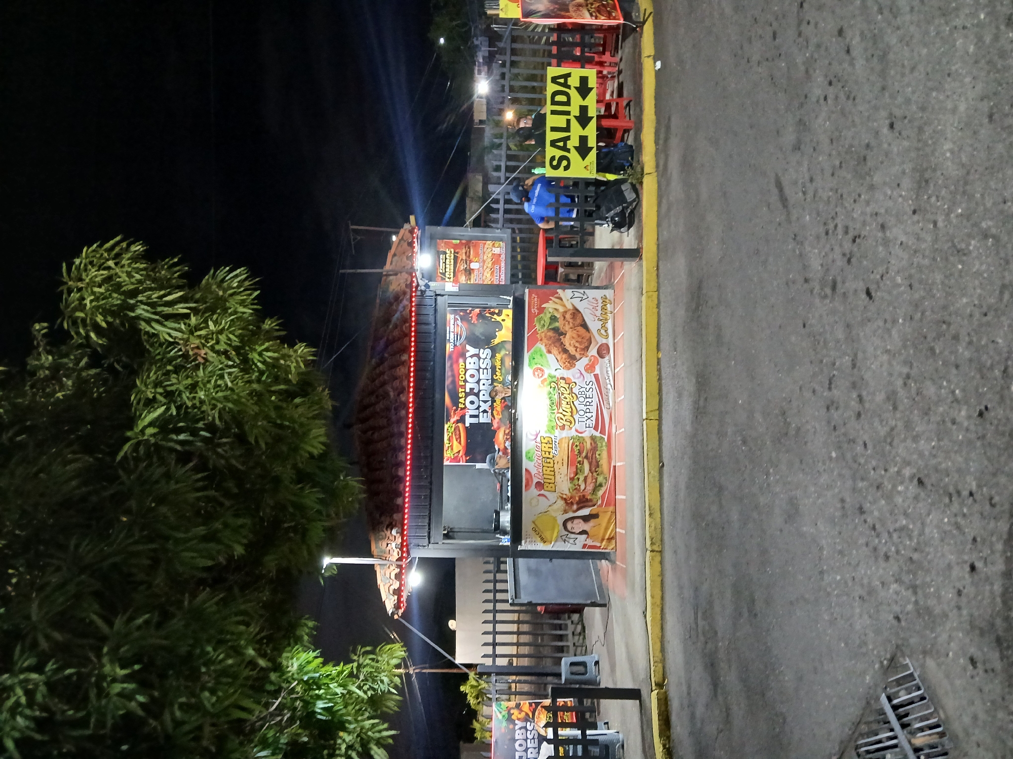 The image displays a nighttime scene in Ciudad Guayana, Venezuela, centered on an illuminated outdoor fast-food stand. The stand, named "Tio Joby Express," is a black structure with a reddish-brown tiled roof, adorned with red LED strip lighting along its perimeter. Promotional banners on the stand depict images of burgers, fried chicken ("Pollo Crujiente"), and a smiling woman. Two individuals are partially visible inside the stand, appearing to be staff. In front of the stand, behind a black metal picket fence, a seating area with red plastic tables and stools is visible. One individual, wearing a blue shirt and cap, stands with their back to the viewer near a yellow sign that reads "SALIDA" (Exit) with two directional arrows. A black backpack and another bag are on the ground beside them. Another person is seated at a table further back. Overhead, dense green foliage from large trees occupies the upper left quadrant of the frame. The foreground consists of asphalt pavement, delineated by a yellow-painted curb. A metal storm drain grate is visible in the lower left of the pavement. A street lamp on a pole provides bright white illumination behind the fast-food stand. The sky is dark, indicating night.
