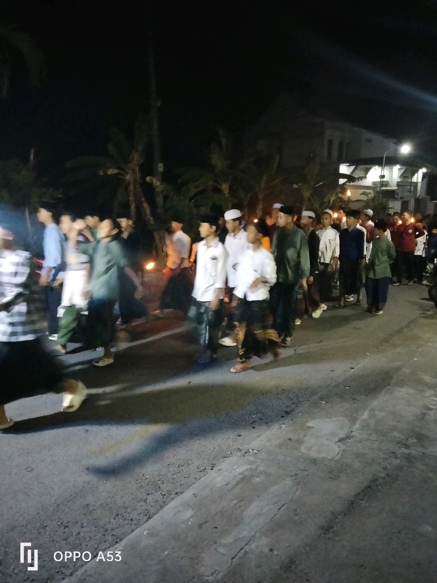 In Bendorejo, Indonesia, at night, a group of young men and boys, most wearing white peci caps, walk in a procession on a paved road. Some are holding torches, illuminating the scene with a warm glow. The participants are dressed in traditional clothing, including long white shirts and sarongs. The background features palm trees and buildings, with a street lamp casting light on the road. The image is captured from a slightly low angle, with the OPPO A53 logo visible in the lower left corner.