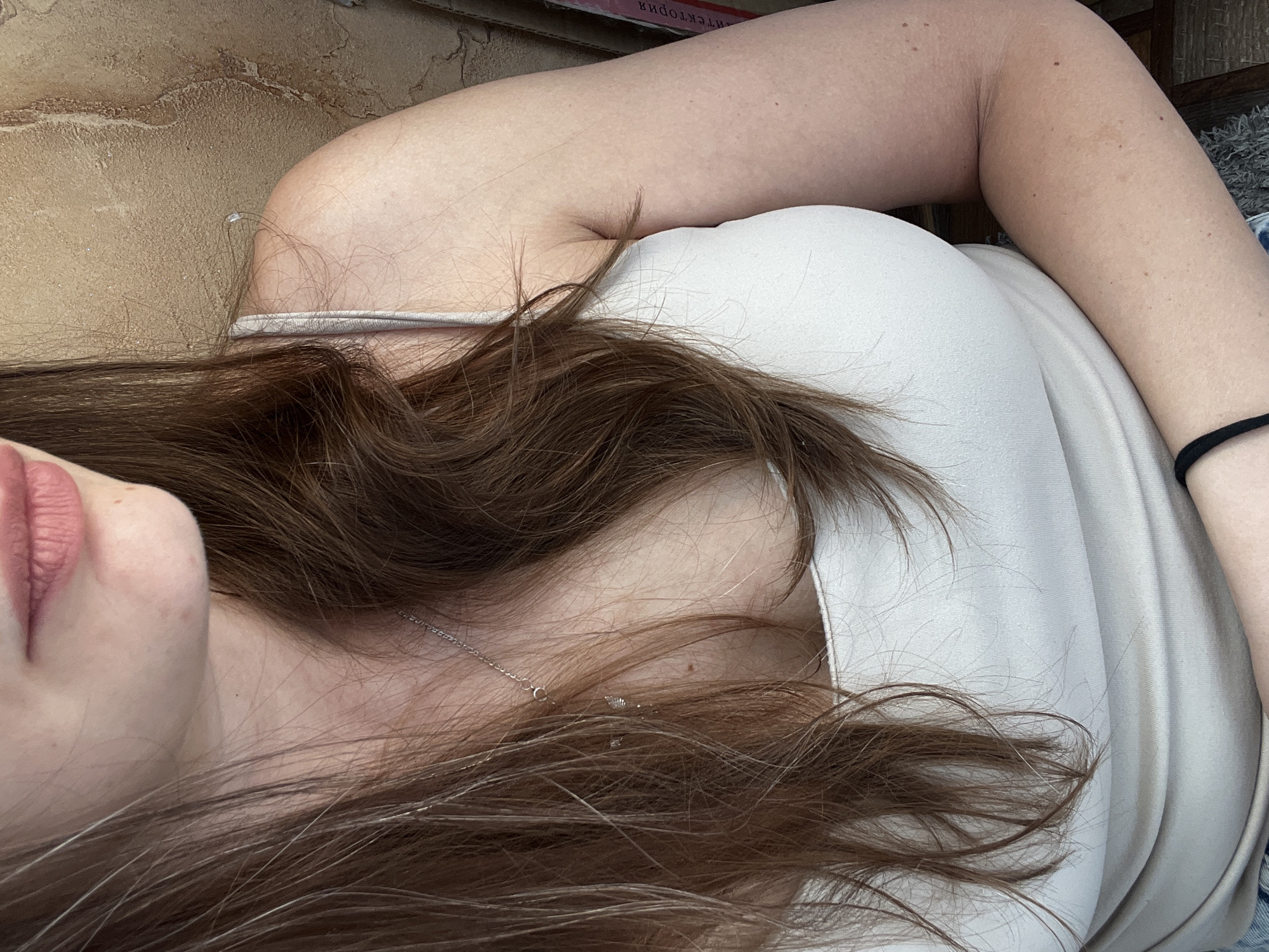 A close-up, waist-up, indoor photograph shows a young woman with long, wavy, light brown hair, wearing a light-colored tank top and a thin silver necklace. Her hair partially obscures her face and torso. The background is a textured, tan-colored wall. The lighting is soft, and the overall scene is calm and casual. The image was captured in the city of Tolyatti, Russia.