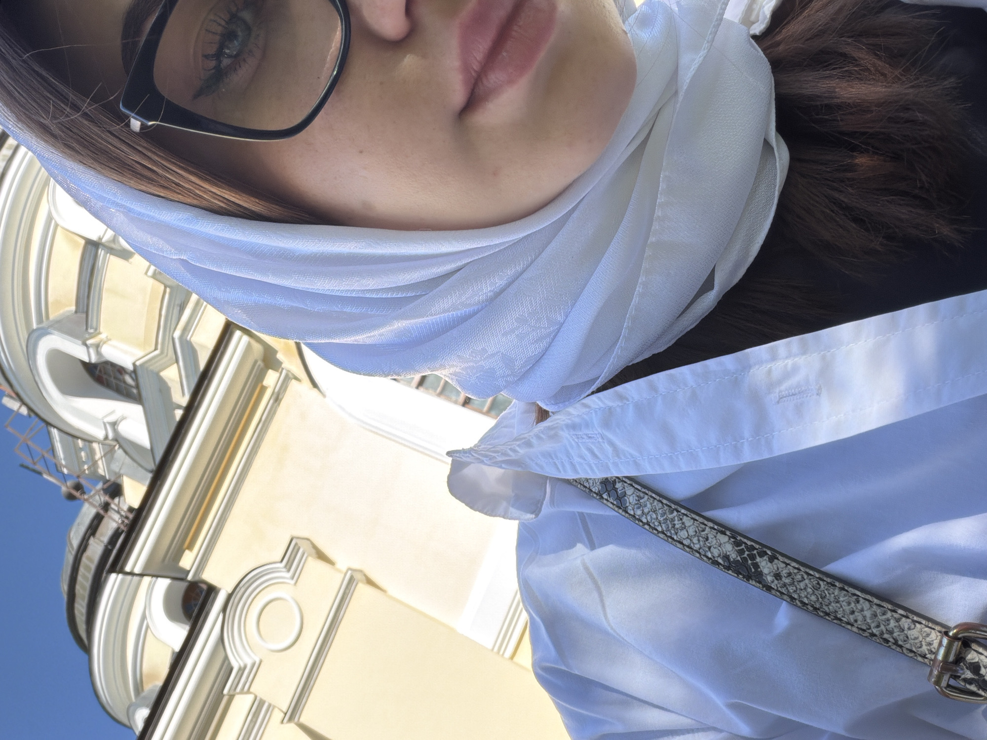 An outdoor, medium shot selfie taken in Căpriana, Moldova, shows a young woman looking at the camera. She is wearing a white headscarf, black-rimmed glasses, and a white collared shirt. A portion of a light yellow building with domes, characteristic of local religious architecture, is visible in the background. The woman's right eye, nose, and lips are visible, along with a strap of her bag. The sky is clear and blue.