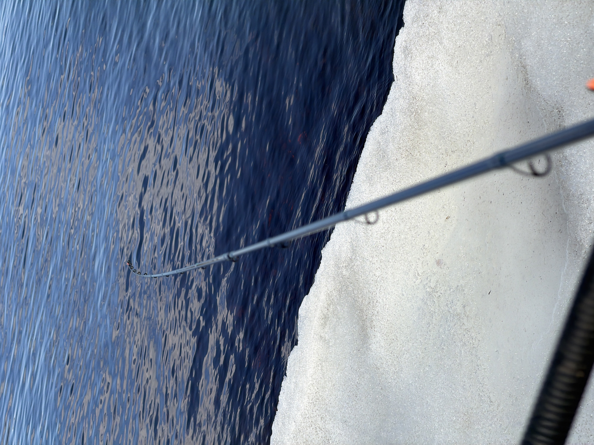 The image shows a close-up view of a fishing rod extending over water, adjacent to a snow-covered surface. The water is dark and rippled, reflecting light. The fishing rod is bent, indicating tension, and the end of the rod is submerged in the water. The snow-covered surface is bright and textured. A portion of what looks like a black, corrugated tube or hose is visible in the lower right corner. The specific location cannot be determined from the image.