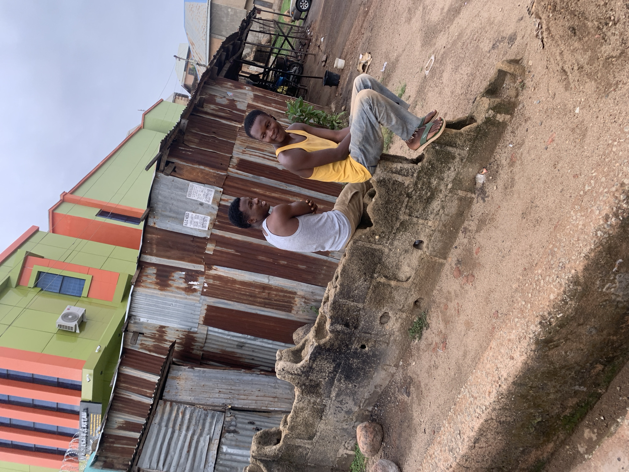 Two young men are sitting on a concrete structure in Jos, Nigeria. One man is wearing a yellow tank top, the other a gray tank top. They appear to be talking. Behind them is a corrugated metal structure and a taller, more modern building painted green and orange. The ground is sandy and uneven. A small structure with metal framing is visible in the background. The sky is overcast.