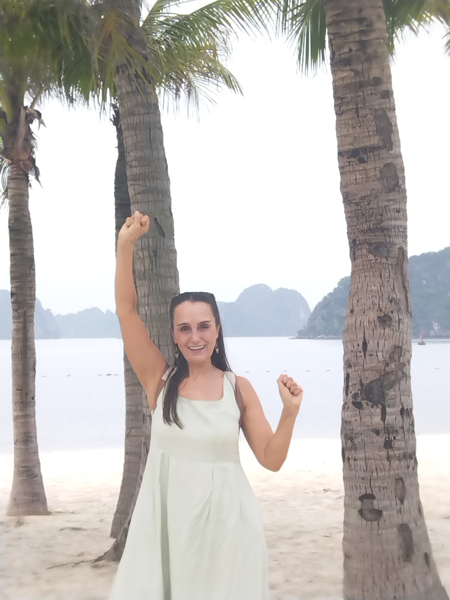A woman stands on a sandy beach, seemingly celebrating with a raised fist. She wears a light-colored dress. Behind her are three palm trees and a body of water with rock formations in the distance. The location is Ha Long Ward, Vietnam.