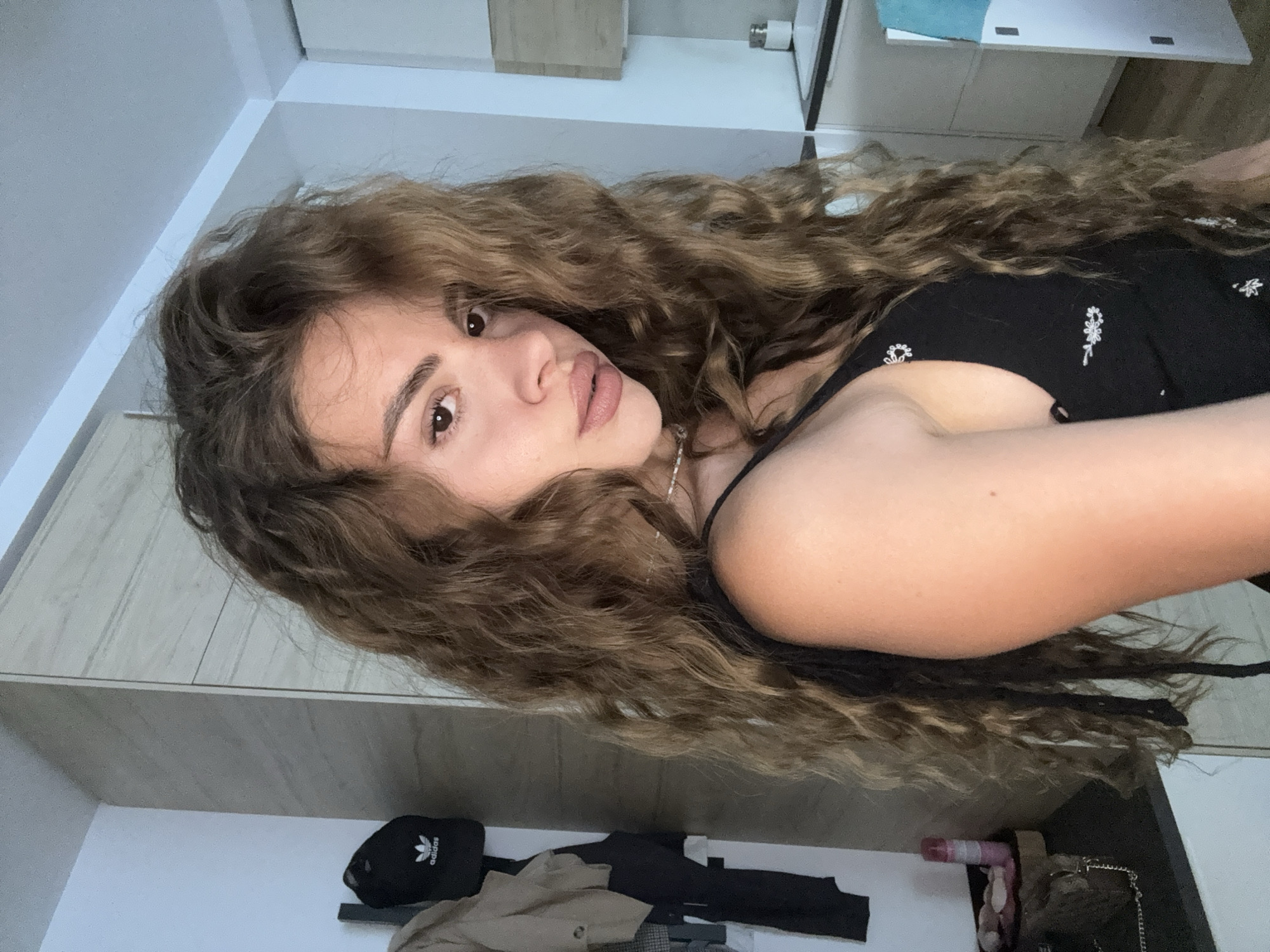 A woman with long, curly brown hair is lying down and looking at the camera. She is wearing a black tank top with small white floral patterns. In the background, there is a white wall with light-colored cabinets and drawers. On the floor below, there is a black Adidas cap and several folded garments. The location is a room in the city of Unknown, Unknown.
