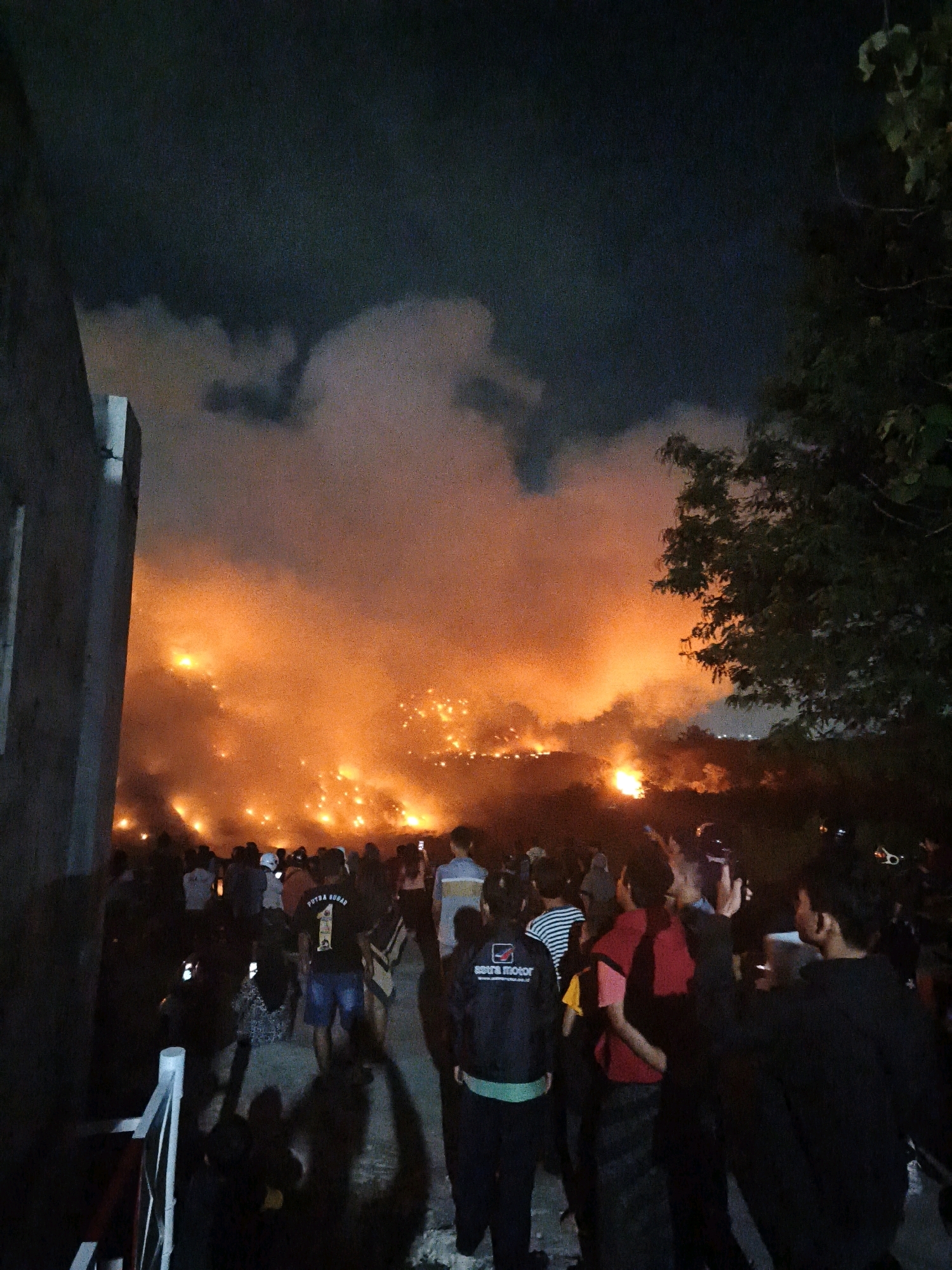 A large crowd of people are gathered at night to witness a significant fire burning in a distant hillside area. The fire illuminates the smoke and clouds above with a bright orange and yellow glow, creating a dramatic and intense scene. Many individuals in the foreground are holding up their phones, likely to record or photograph the event. The presence of a building structure on the left side of the image suggests an urban or semi-urban environment. The overall atmosphere appears to be one of awe and concern. This event took place in the city of Surakarta, Indonesia.