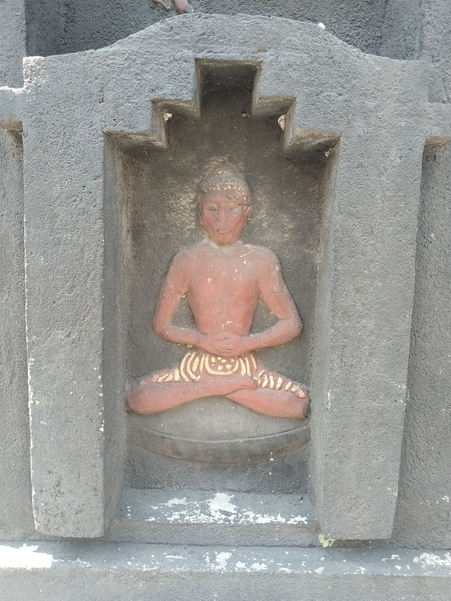 The image depicts a carved statue of a seated figure in a niche. The figure is reddish-brown in color, with its legs crossed in a lotus position and hands resting in its lap. There are decorative markings around the waist and legs. The niche is gray and textured, with a stepped design at the top. The statue appears to be weathered, and there is some discoloration at the base of the niche. Based on the provided information, the location is Masaran, Indonesia.