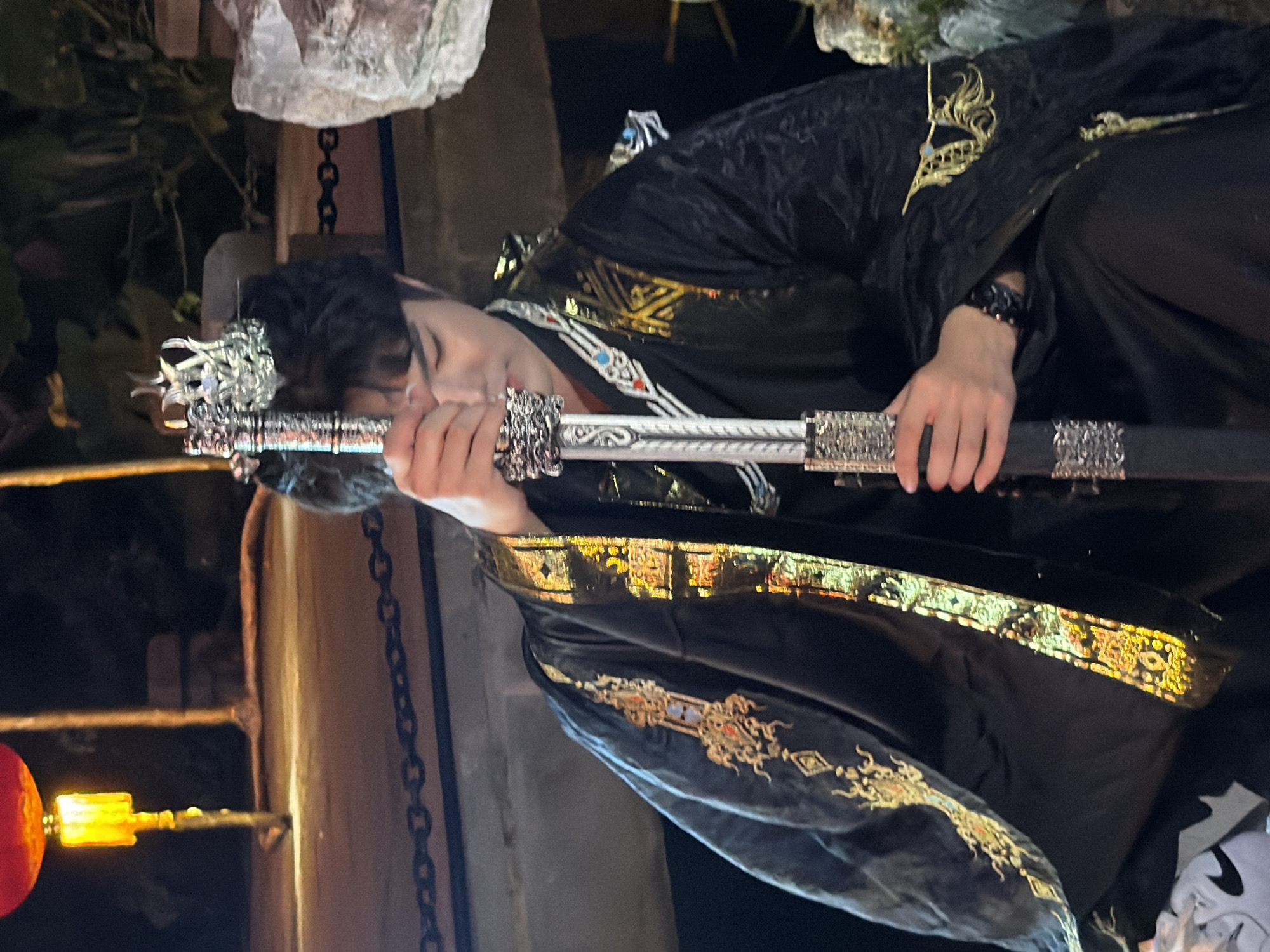 The image shows a person in a black, ornate robe with gold embroidery, holding a sword. The person appears to be male, with dark hair and a crown on their head. They are lying down, with their head resting on their arms, which are holding the sword. The sword has a decorative hilt and scabbard. The background is dark and appears to be outdoors, with some foliage visible. There is also a red lantern with a yellow light hanging in the lower left corner of the image. The scene has a dramatic and somewhat somber mood, possibly depicting a character in a historical or fantasy setting. The time of day is likely evening or night, given the lighting conditions and the presence of the lantern. The person's eyes are closed, suggesting a state of rest or contemplation. The intricate details of the robe and sword suggest a high-status or important character. The overall composition and subject matter hint at a narrative or storytelling element. There is no visible text in the image.