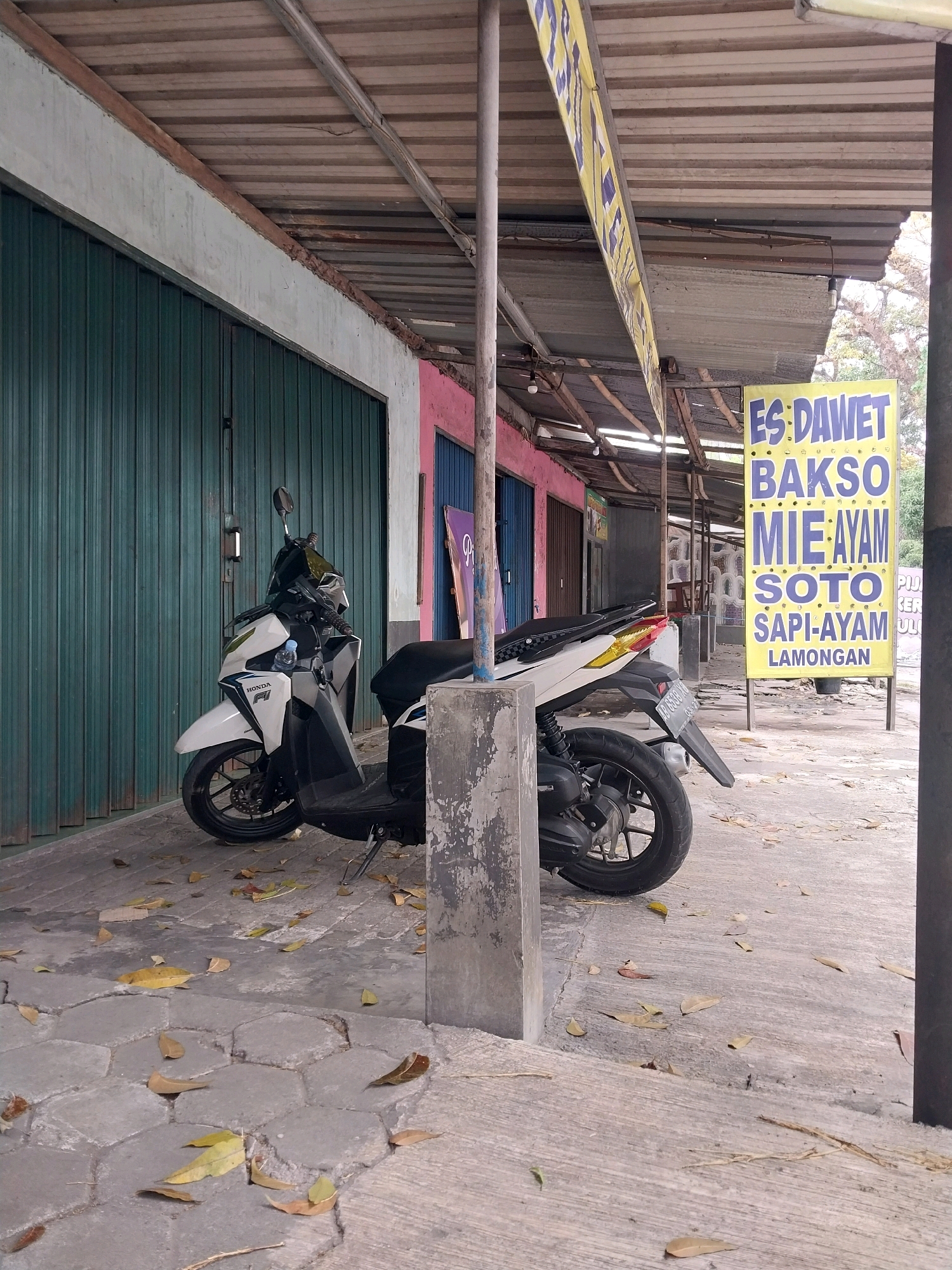 The image shows a street scene in Tamanmartani, Indonesia. A white and black scooter is parked on a sidewalk in front of a row of shops with green metal roll-up doors. A pink building is visible behind the scooter. To the right, a yellow sign with blue text advertises "ES DA WET BAKSO MIE AYAM SOTO SAPI-AYAM LAMONGAN". The sidewalk is paved with hexagonal grey stones and scattered with fallen leaves. A concrete pillar is in the foreground, partially obscuring the scooter. The shops have a corrugated metal roof supported by wooden beams and metal poles. The overall environment appears to be an outdoor commercial area.