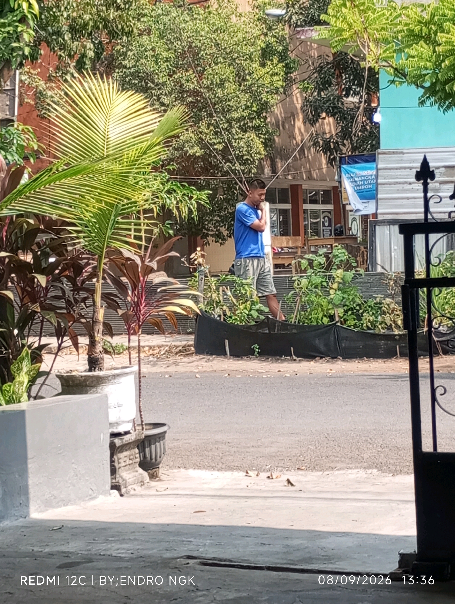 The image shows a man in a blue t-shirt and grey shorts standing outdoors in front of a building. He appears to be looking down at his phone. There are trees and plants in the foreground and background. The scene is well-lit by daylight. A text overlay at the bottom of the image reads "REDMI 12C | BY:ENDRO NGK" and "08/09/2026 13:36". The context of Nganjuk, Indonesia is not discernible from the image content alone.
