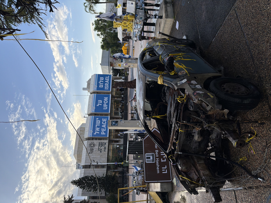 The image depicts Rabin Square in Tel Aviv-Yafo, Israel, facing the Tel Aviv-Yafo Municipality building. In the immediate foreground, a heavily damaged, dark-colored four-door sedan is prominently displayed. The car's front end is completely destroyed, with charred engine components and twisted metal exposed. The interior is gutted and blackened. Numerous yellow ribbons are tied to various parts of the vehicle, including the steering column, door frames, and exterior. The car rests on a damp, paved surface.
To the left of the vehicle, a brown sign with white Hebrew text "הר בית השם" and a graphic is partially visible. To the right, a makeshift memorial setup includes a series of vertical elements adorned with Israeli flags, yellow ribbons, and other objects. Further right, a decorated structure, possibly a piano, is also covered in yellow ribbons and various items. Additional yellow ribbons are suspended from an unseen overhead structure or tree branch.
In the background stands the Tel Aviv-Yafo Municipality building, a large, light-colored, multi-story structure. Its facade features three prominent blue banners, collectively displaying "PEACE UPON ISRAEL" in both English and Hebrew ("שלום על ישראל"). Individuals are discernible through the building's large windows. Tall trees, including a conifer on the left and palm trees on the right, are visible. The sky is partly cloudy with patches of blue. Thin, dark lines, resembling wires or cables, traverse the upper portion of the frame.