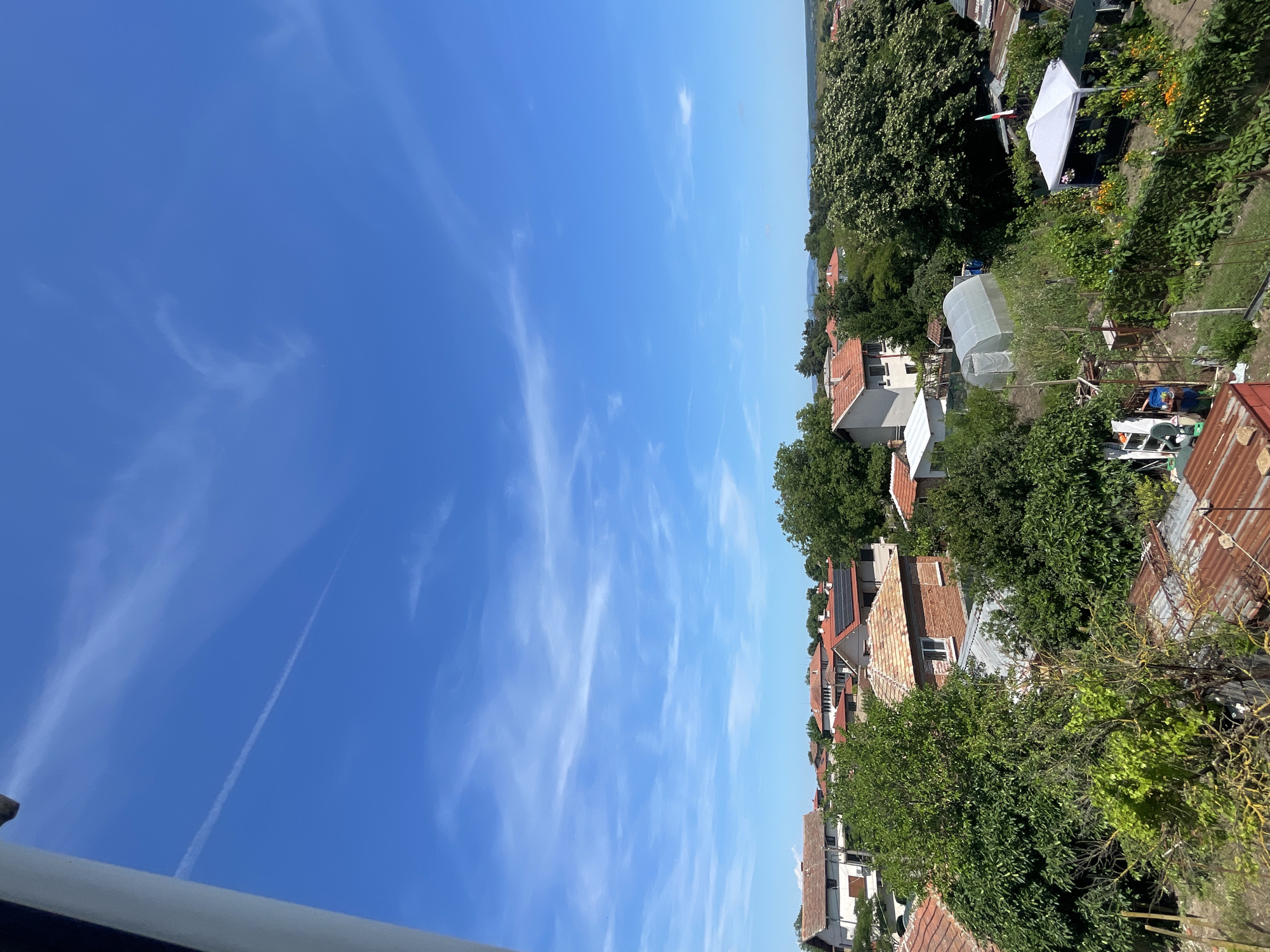 The image shows a view from an elevated position looking over a residential area in the city of Debelt, Bulgaria, under a clear blue sky with scattered wispy clouds and a visible contrail. In the foreground, a white railing is partially visible on the left. The scene is characterized by a cluster of houses with red-tiled roofs, interspersed with lush green trees and vegetation. Several structures are visible, including a greenhouse and a white pop-up canopy tent in a garden area. The overall impression is a sunny day in a semi-rural or suburban setting.