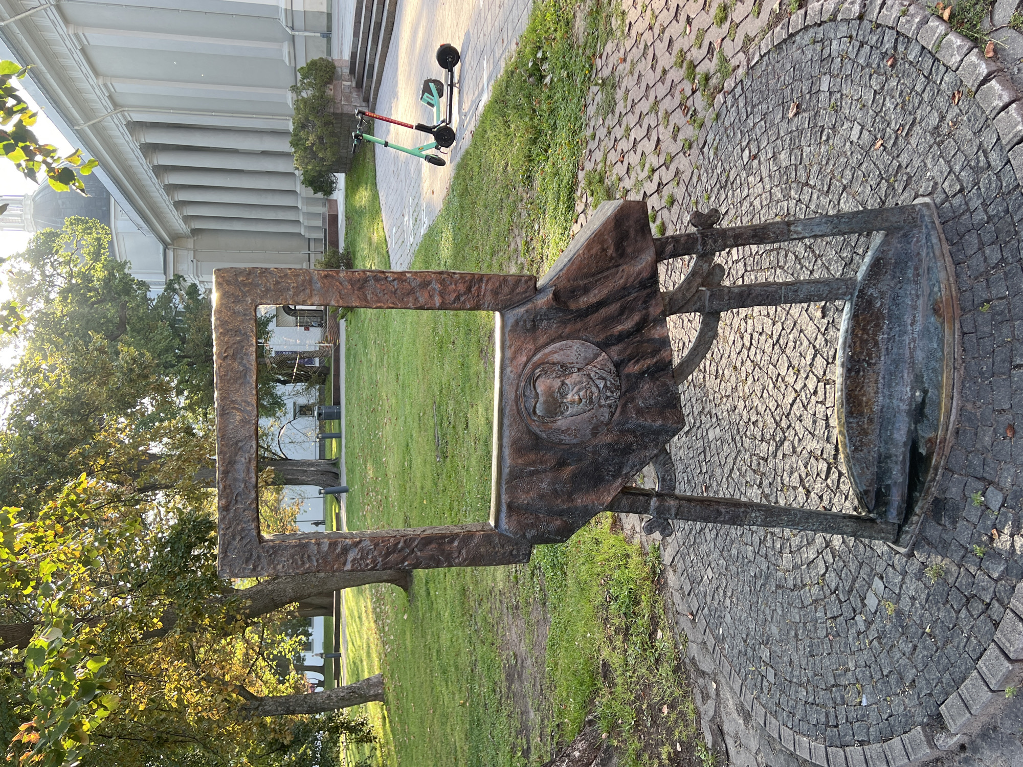 The image depicts an outdoor scene in Vilnius, Lithuania, likely on a bright, sunny day. In the foreground, a large, weathered metal sculpture resembling an easel frames an empty space, suggesting a placeholder for a painting or a person. The sculpture is ornately decorated, featuring a relief of a man's portrait on its base. It stands on a circular paved area made of cobblestones.
Behind the sculpture, a well-maintained park-like area unfolds. Lush green grass covers the ground, interspersed with fallen autumn leaves. Several trees with vibrant green and yellow foliage are visible, casting shadows on the grass. In the middle ground, a pathway made of light-colored paving stones leads towards a grand building with a colonnaded facade and a domed structure, possibly a significant architectural landmark in Vilnius. Two electric scooters, one green and one red, are parked on this pathway. The background is dominated by the imposing architecture of the building and the natural beauty of the park. The overall atmosphere is serene and picturesque, capturing a moment of calm in an urban setting.