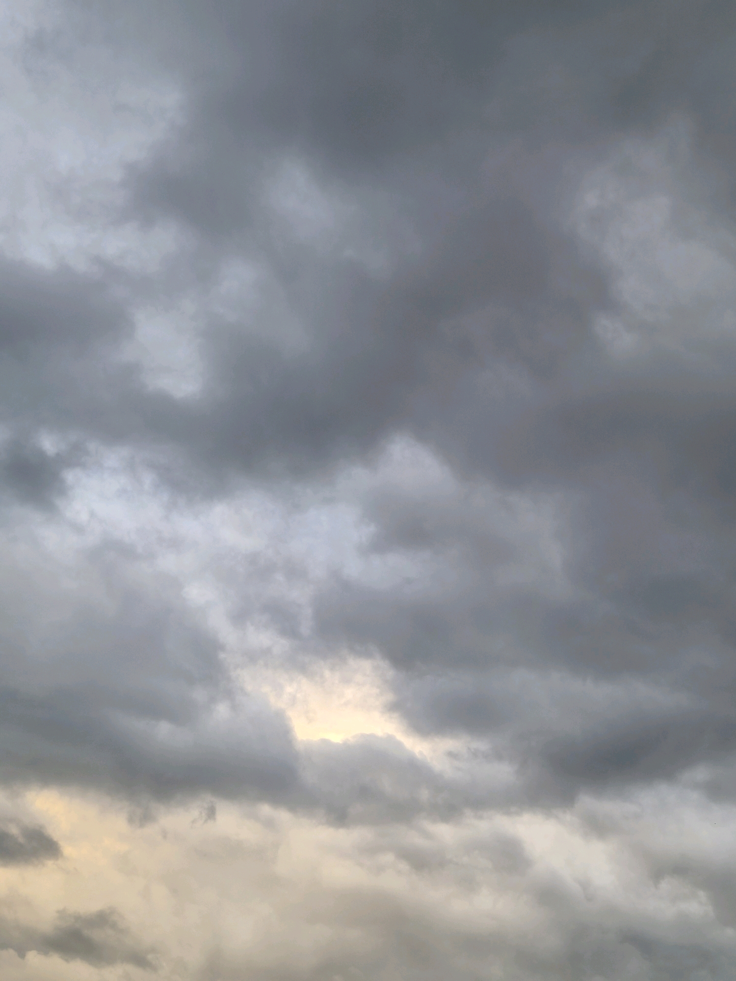 The image depicts a cloudy sky over Schiffdorf, Germany. The clouds are predominantly dark gray, with some lighter patches suggesting a break in the cloud cover where sunlight is attempting to penetrate. The overall atmosphere is somber and overcast. There are no people, subjects, objects, or actions visible in the scene, only the natural phenomenon of the sky. The photograph captures a moment of atmospheric conditions without any specific human or manufactured elements.