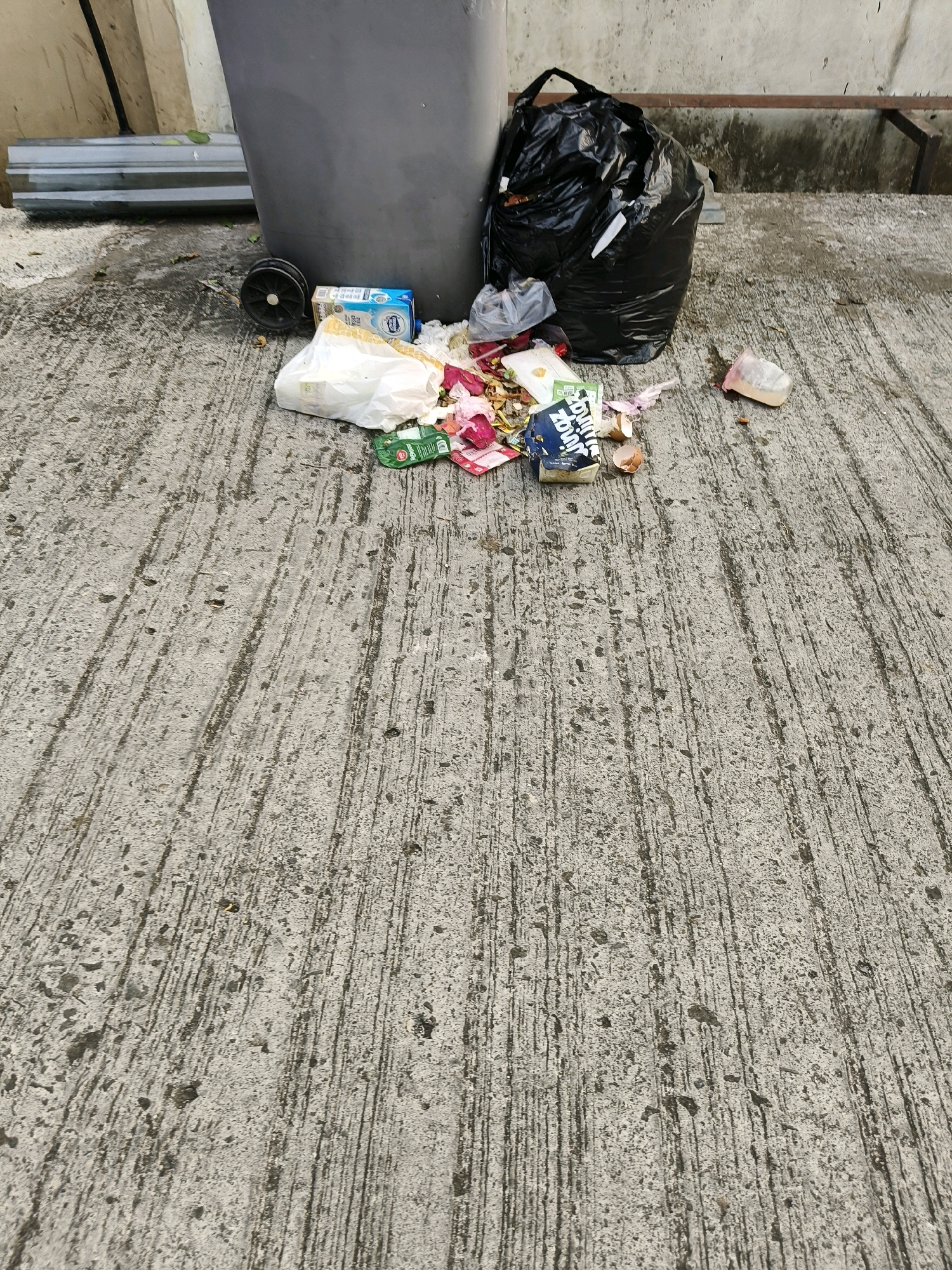 An outdoor scene depicts a collection of trash next to a large grey trash bin on a concrete surface. The trash includes a black plastic bag filled with refuse, a white plastic bag, a blue carton, and scattered food wrappers, eggshells, and pink flower petals. In the background, a beige wall and a metal barrier are visible. The concrete surface is textured with parallel lines. The scene appears to be located in an urban environment, possibly in Bandung City, Indonesia, given the context provided. There are no people visible in the image.